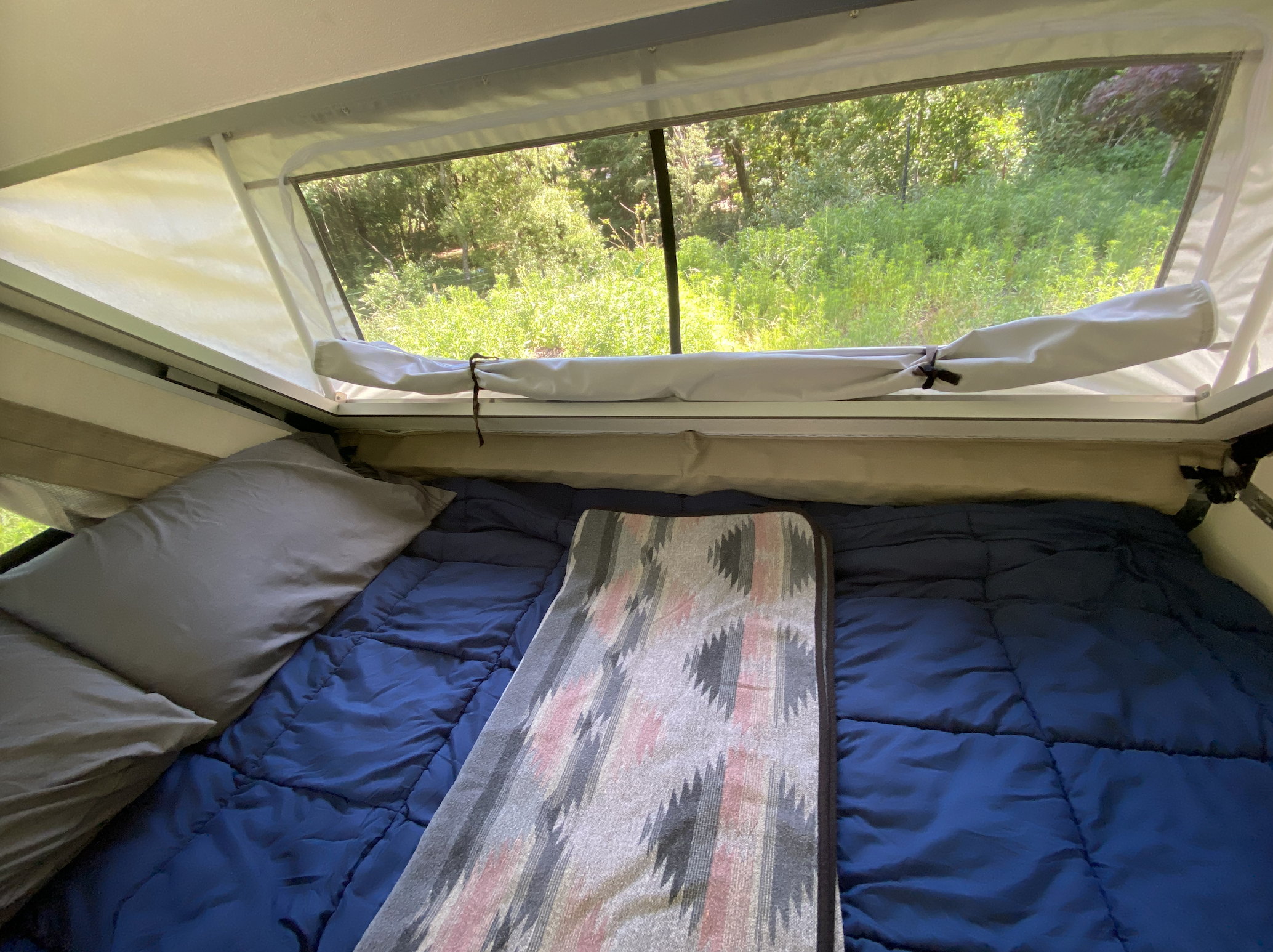 Cozy RV rental: bed with blue comforter & patterned blanket, large window view of vibrant green nature.. A-Liner Classic 2019