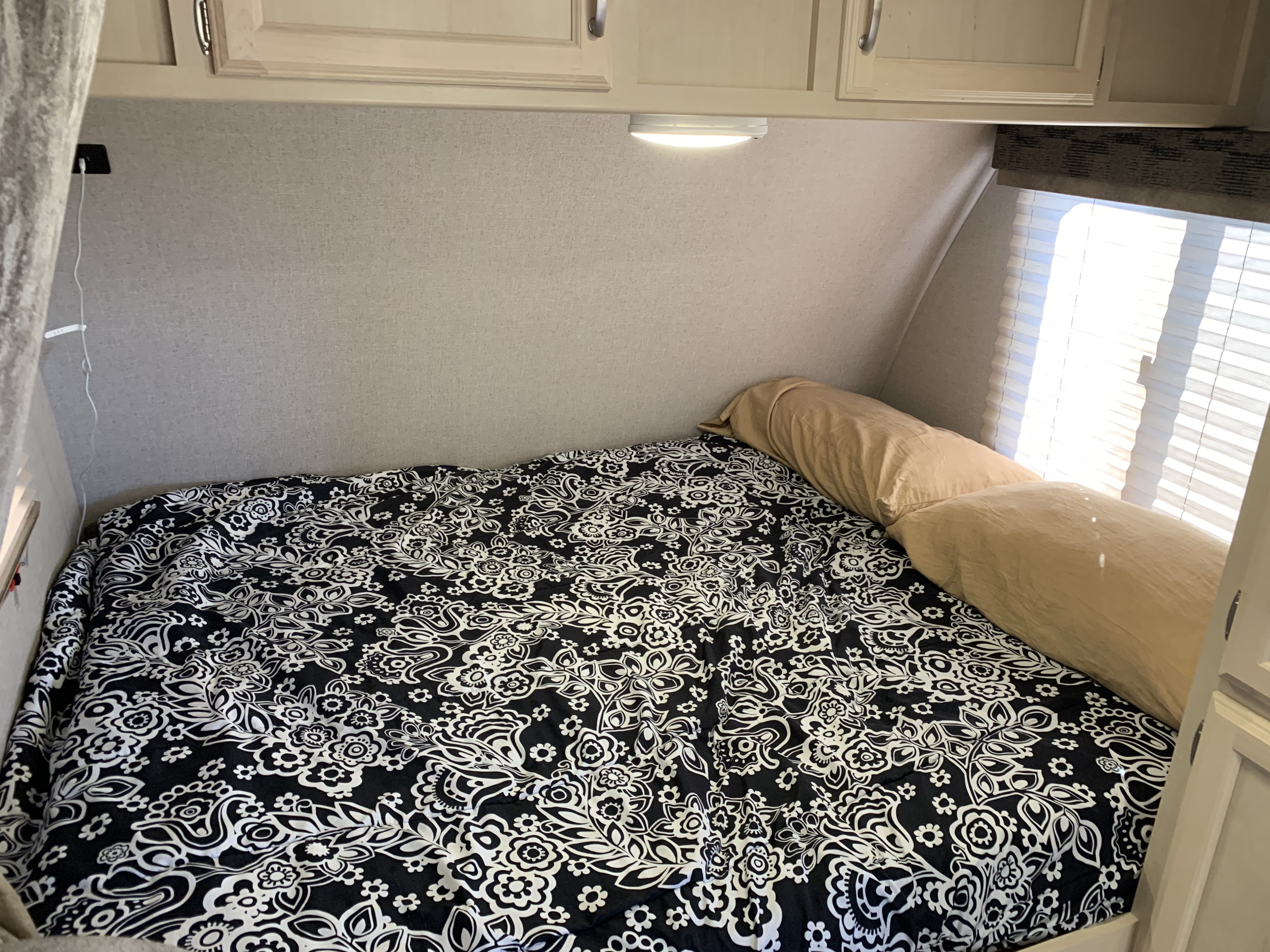 Comfortable RV rental bedroom featuring a patterned bed, tan bolster pillow, cabinets, and charging port. Bright window view.. Other Other 2019