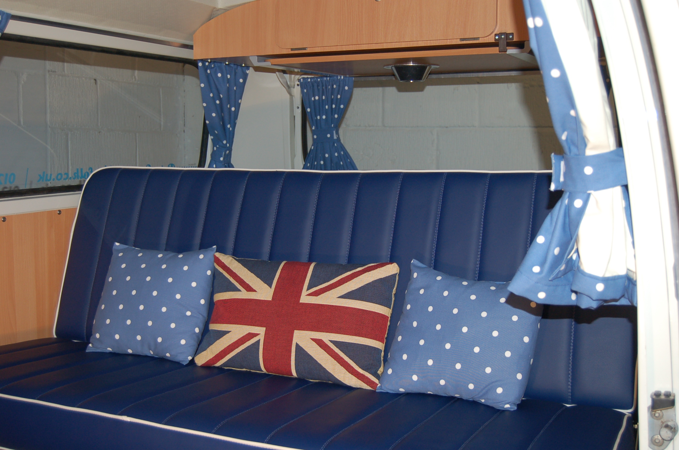 Inviting blue padded seating in an RV rental, complete with polka-dot and Union Jack cushions, & tie-back curtains.. Volkswagen 2.0L T2 Late Bay 1975