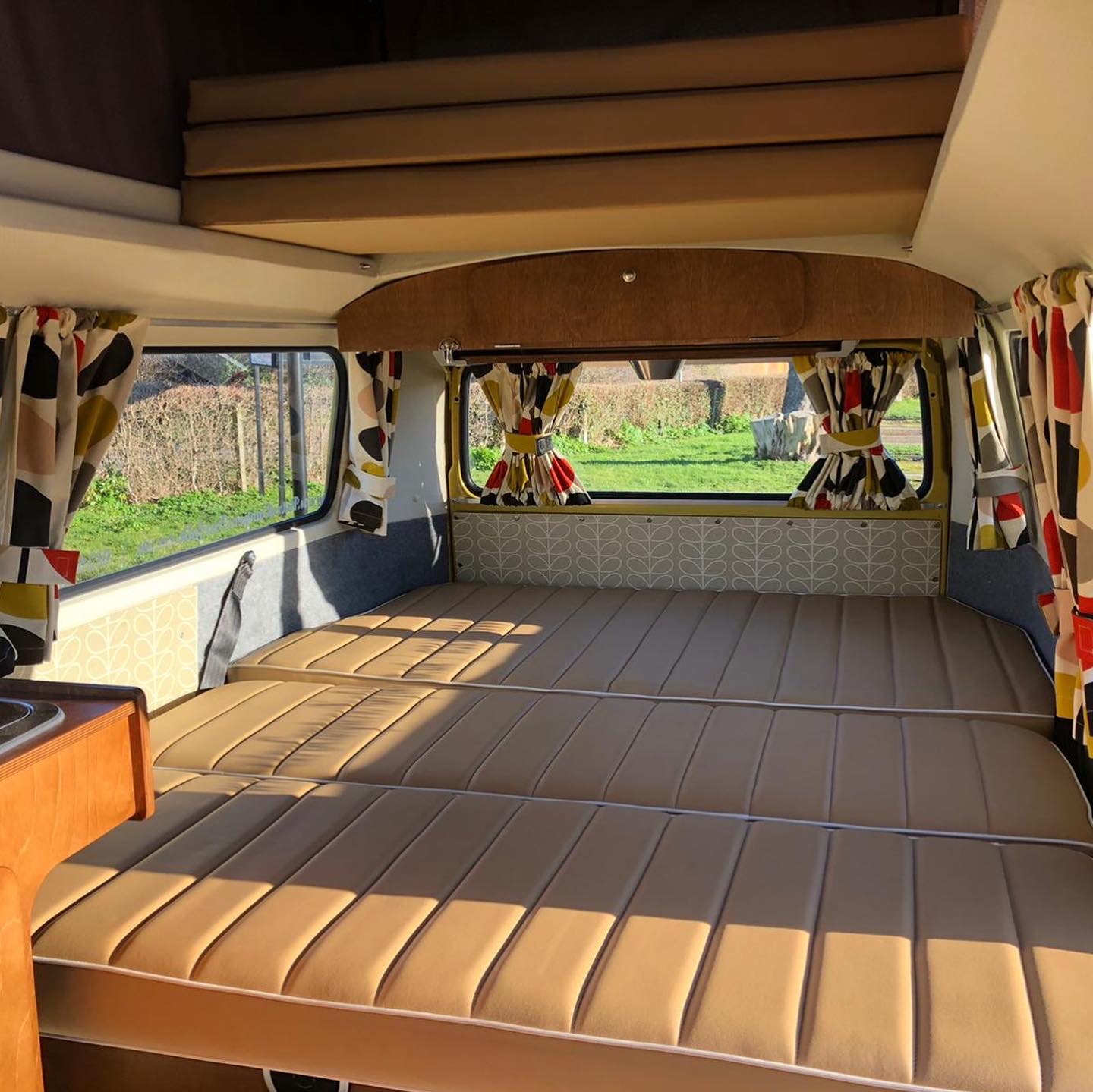 Experience this RV rental: a large tan bed, colorful patterned curtains, and an overhead sleeping area in a bright camper.. Volkswagen 2.0L T2 Late Bay 1975