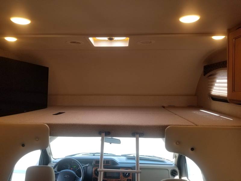 RV rental interior: A cozy overhead bunk bed, driver's cockpit, and TV are illuminated by ceiling lights.. Thor Chateau 2017