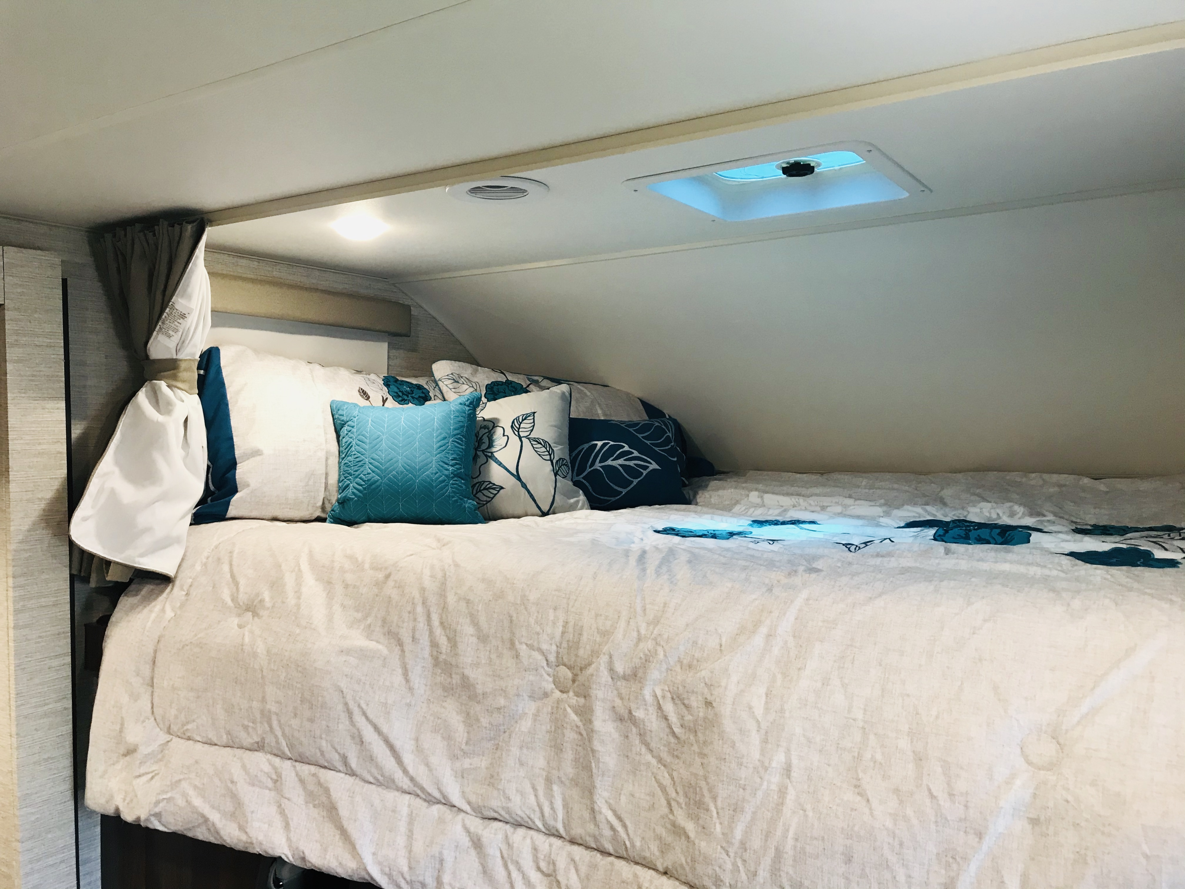 Cozy RV rental over-cab bed with a light comforter, teal pillows, ceiling light, and skylight above for travel.. Winnebago Minnie Winnie 2020