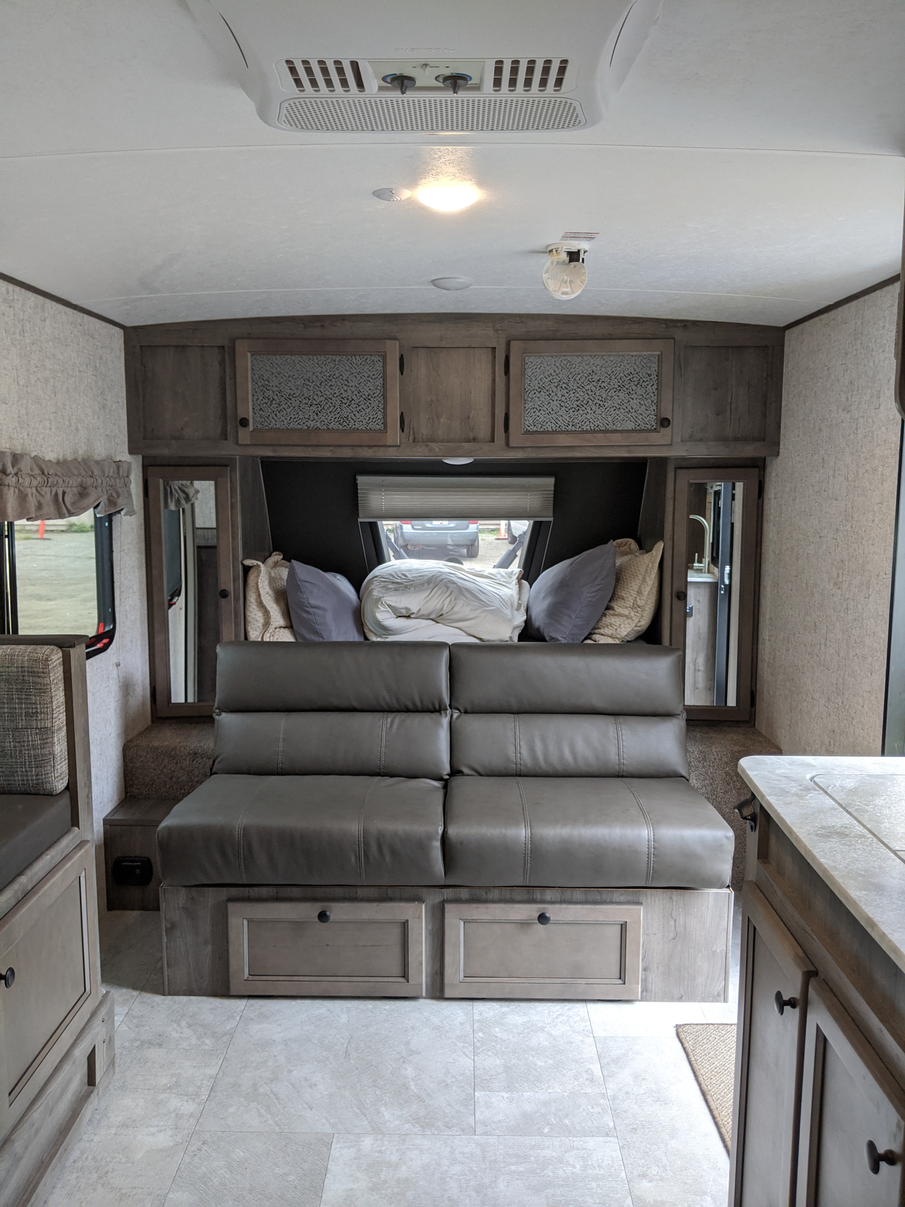 Cozy RV rental interior featuring a grey sofa, overhead cabinets, light floor, and visible AC unit on the ceiling.. Coachmen Apex 2021