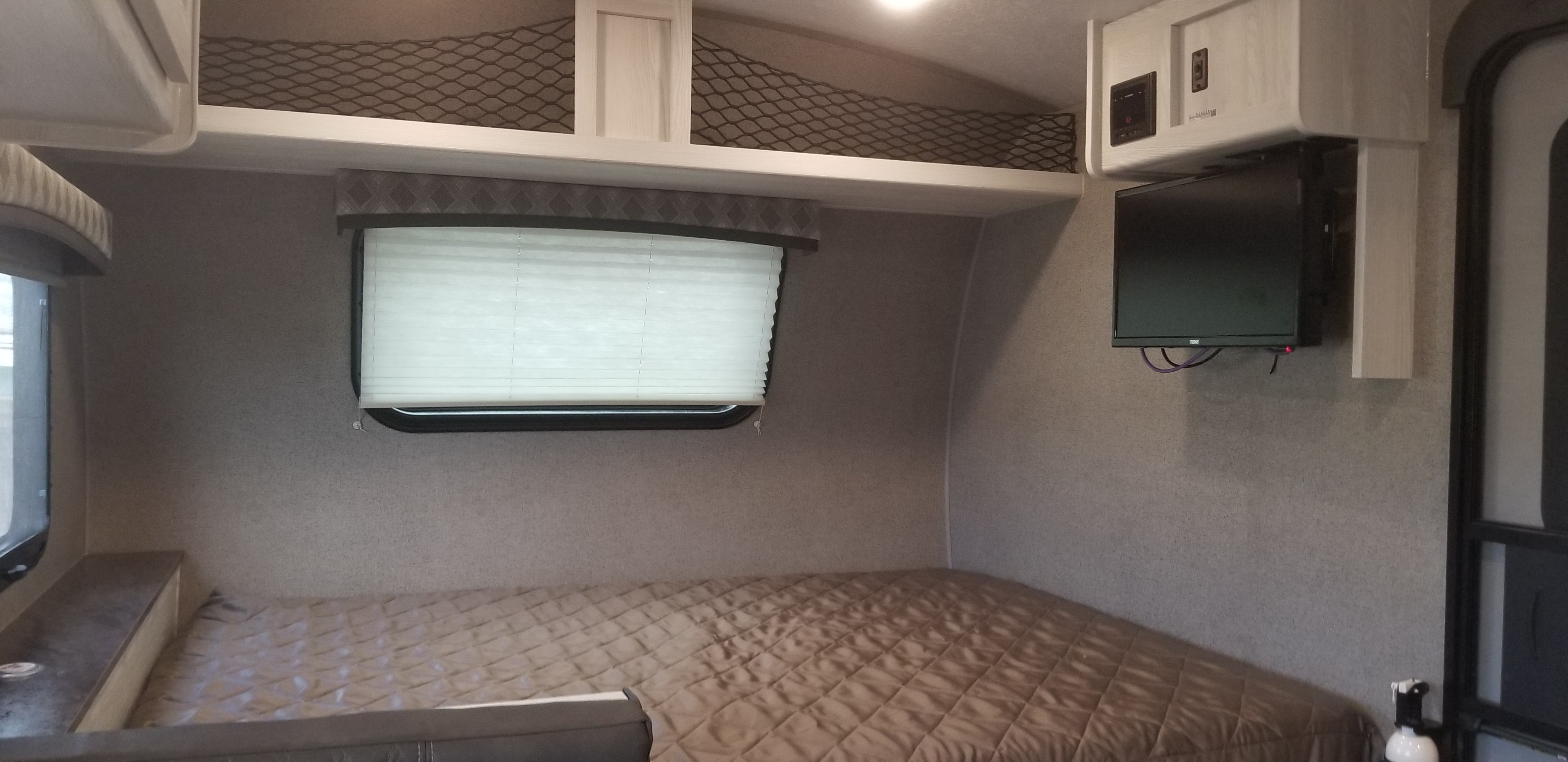 Compact RV rental bedroom showing a bed, window with blinds, wall-mounted TV, and overhead storage netting.. Forest River Flagstaff 2020