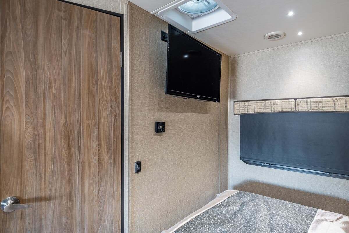 RV rental bedroom: a wood-look door, mounted TV, bed, and window with shade. Modern and compact.. Dynamax Corp Isata 2017