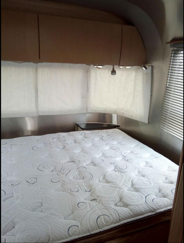 RV rental bedroom: A comfortable sleeping area with a quilted white mattress, overhead cabinets, and window blinds in a shiny camper.. Airstream Safari 2007