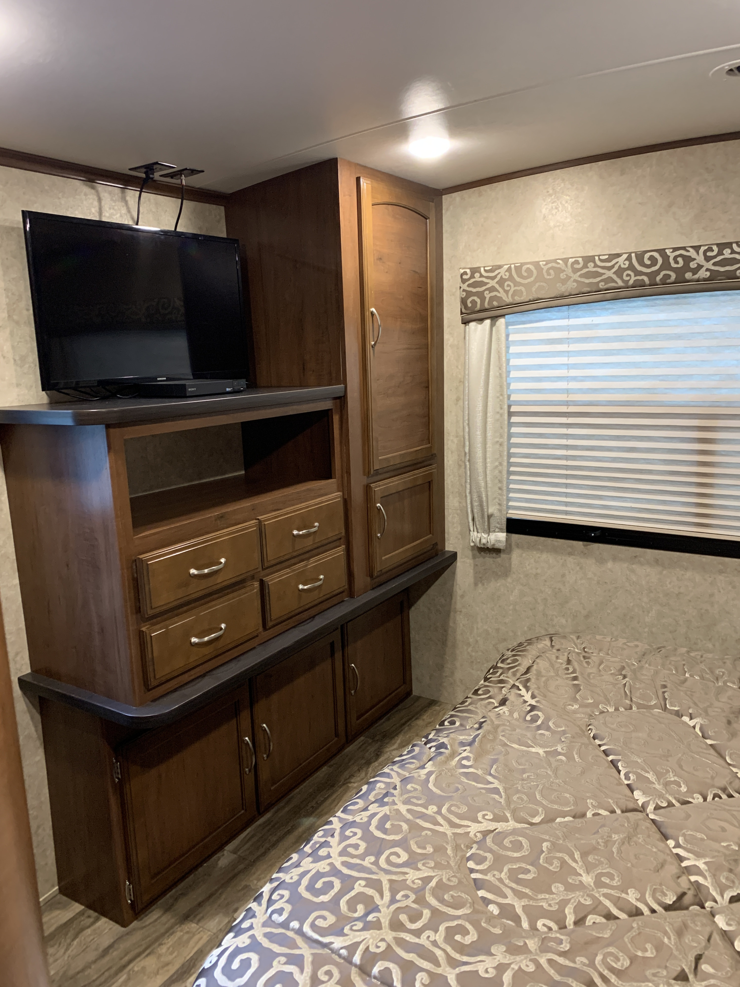 Compact rv rental bedroom: dark wood built-in storage, TV on stand, drawers, tall cupboards, and a bed with patterned linens.. Jayco White Hawk 2017