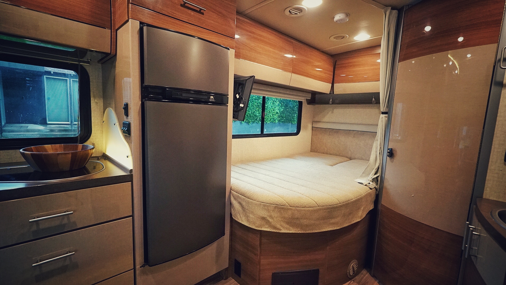 This modern rv rental interior features a compact kitchen with fridge, cozy curved bed, and mounted TV setup.. Mercedes Sprinter 2017