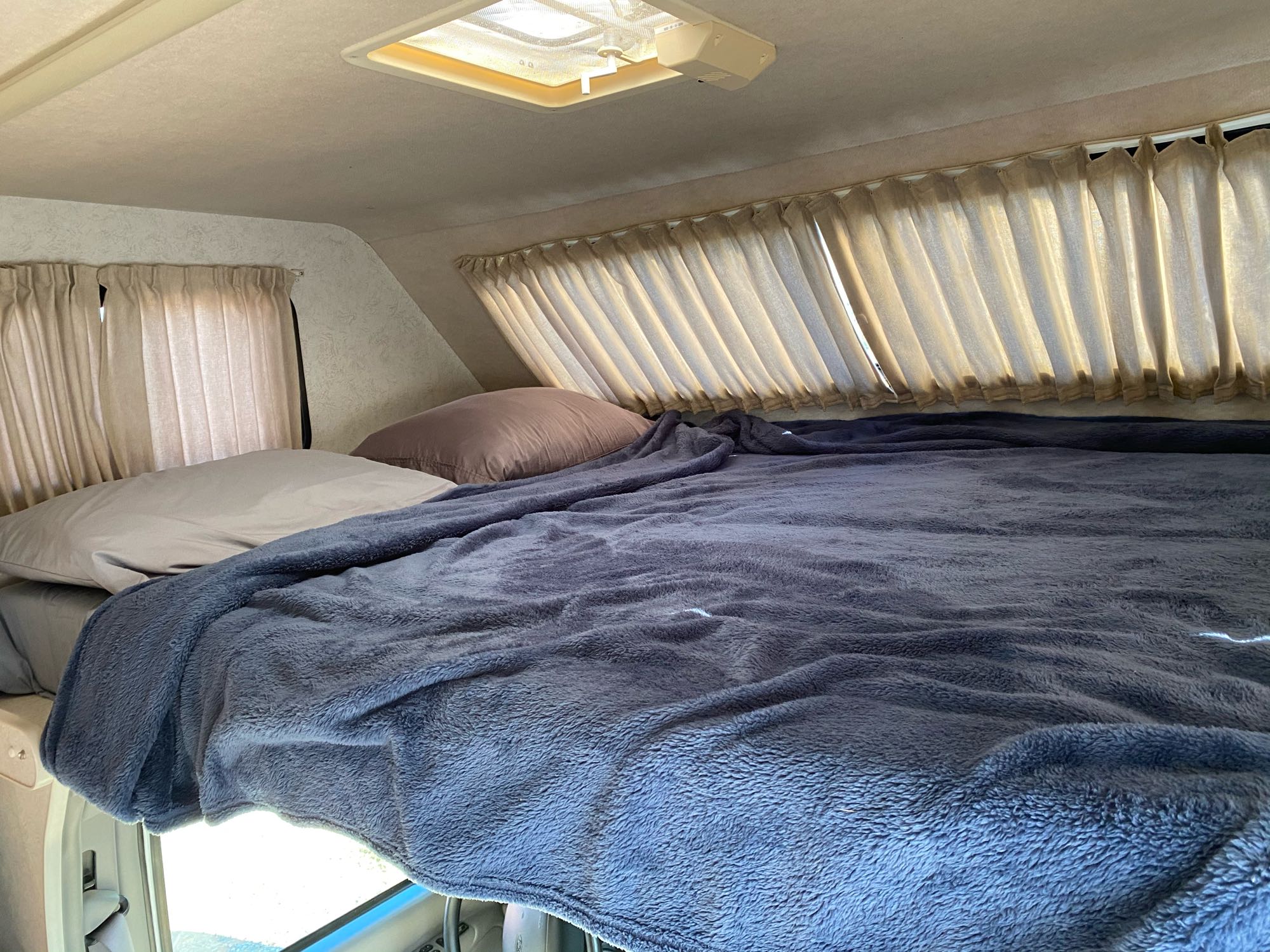 Cozy RV rental sleeping area with a comfortable bed, blue blanket, pillows, curtains, and roof vent.. Itasca Spirit 2000