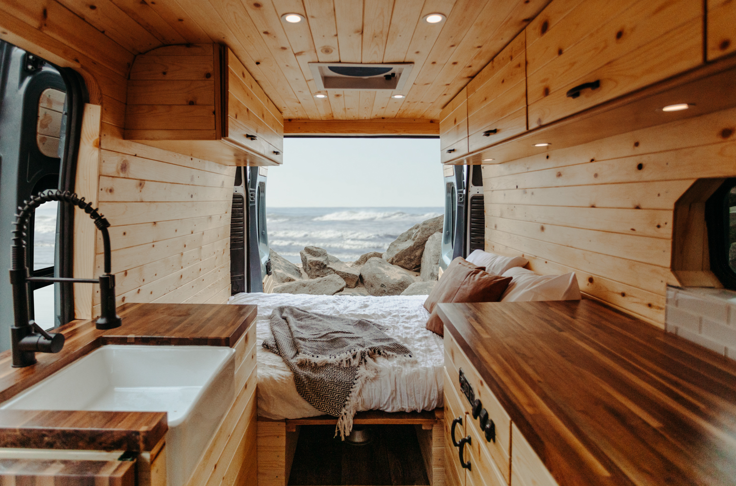 Inside a custom wood-paneled rv rental camper van. Features a kitchen, comfortable bed, and scenic ocean view for mobile living.. Ram Promaster 2019
