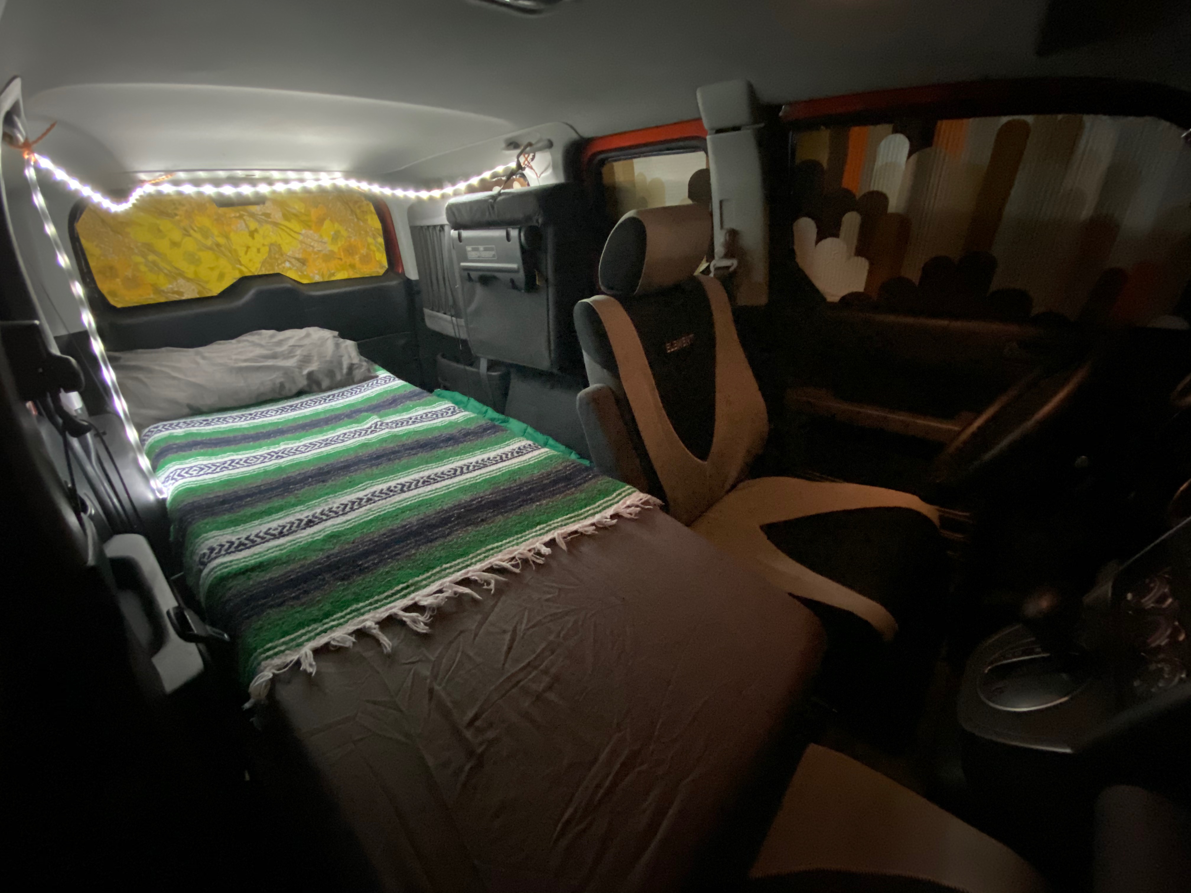 Cozy car camping interior of an RV rental, featuring a bed with a colorful blanket and string lights.. Honda Element 2005
