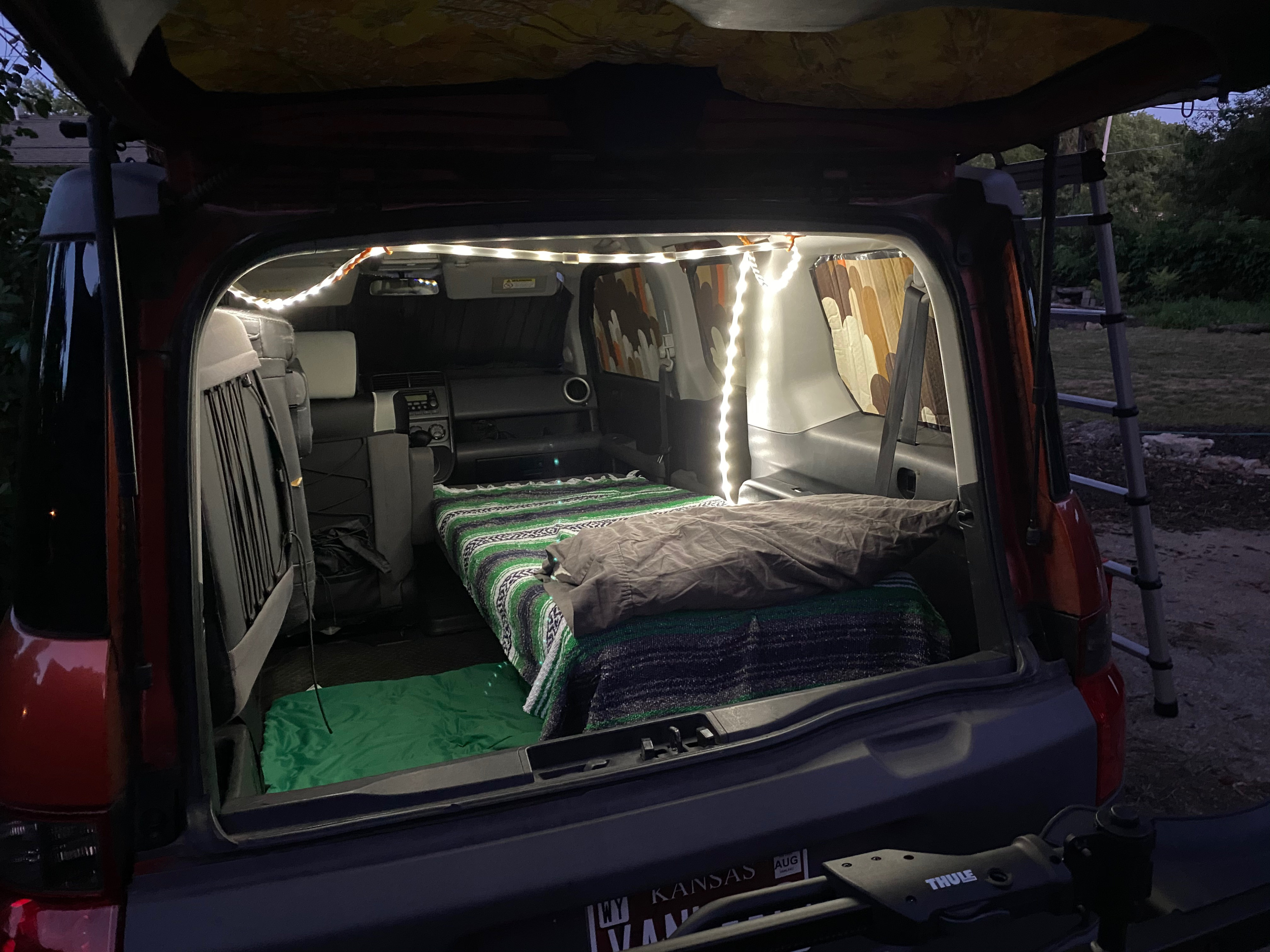 Compact RV rental interior illuminated by LED lights, featuring a cozy bed. Ideal for car camping adventures with a Thule rack visible outside.. Honda Element 2005
