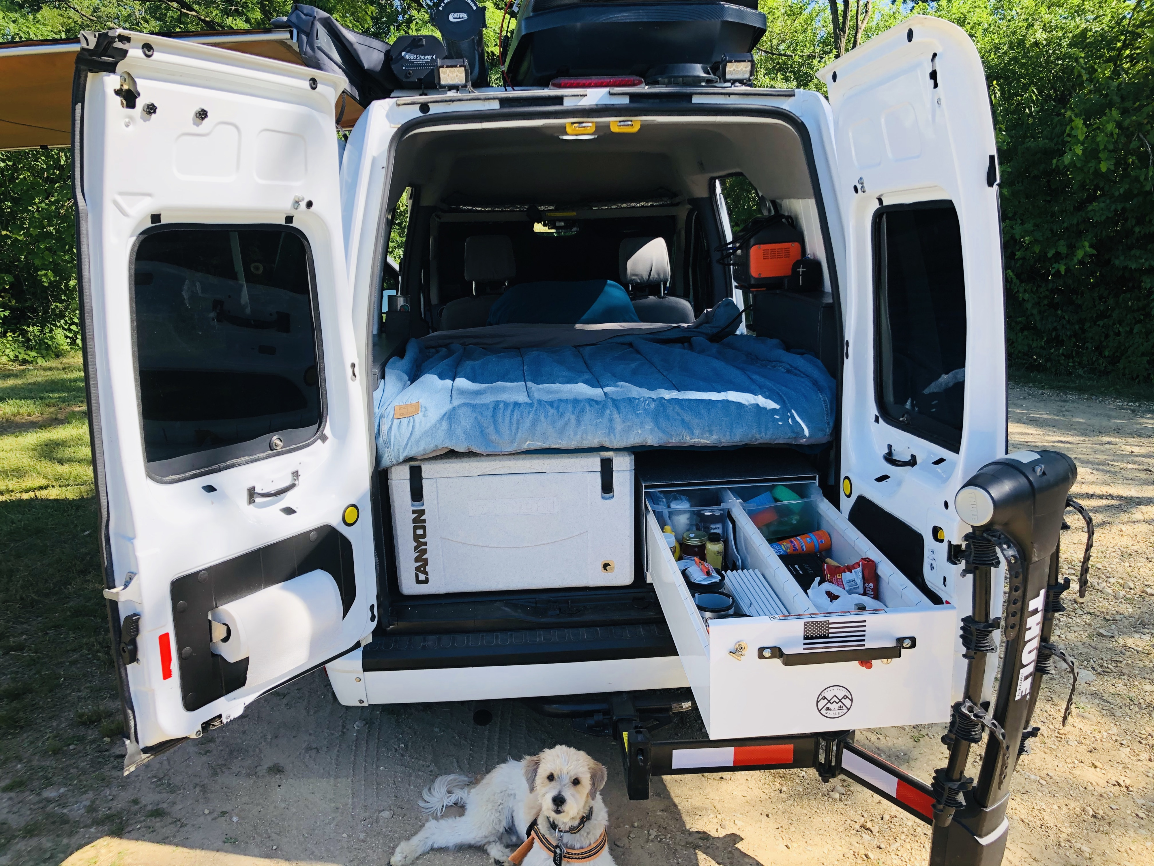Explore with this rv rental! Open rear of a white camper van shows a bed, cooler, pull-out gear storage, and a dog.. Ford Transit 2011