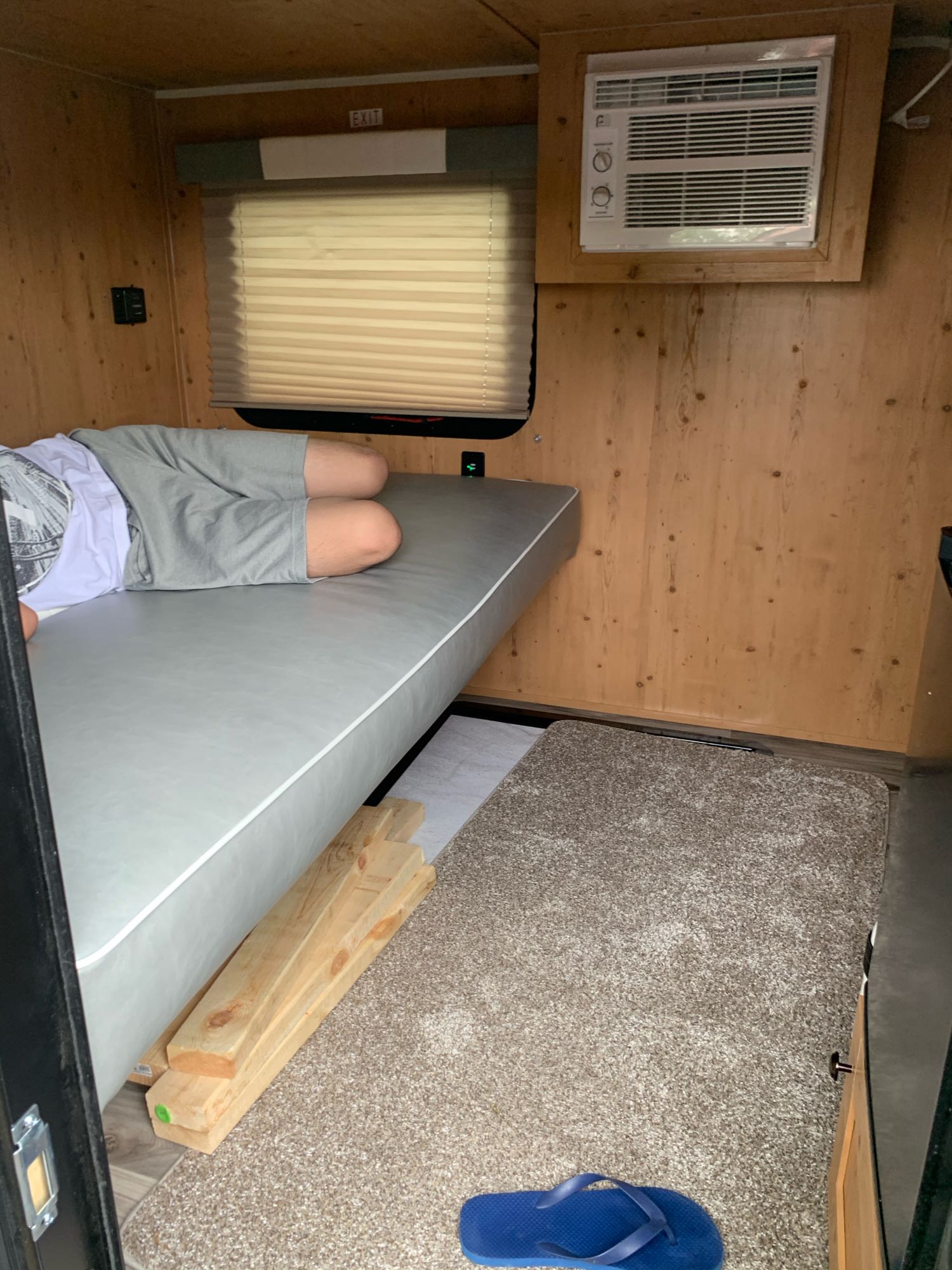Inside an RV rental: a person lying on a gray bunk, with an AC unit, window, and wood-paneled walls.. Sunset Park RV SunRay 2021