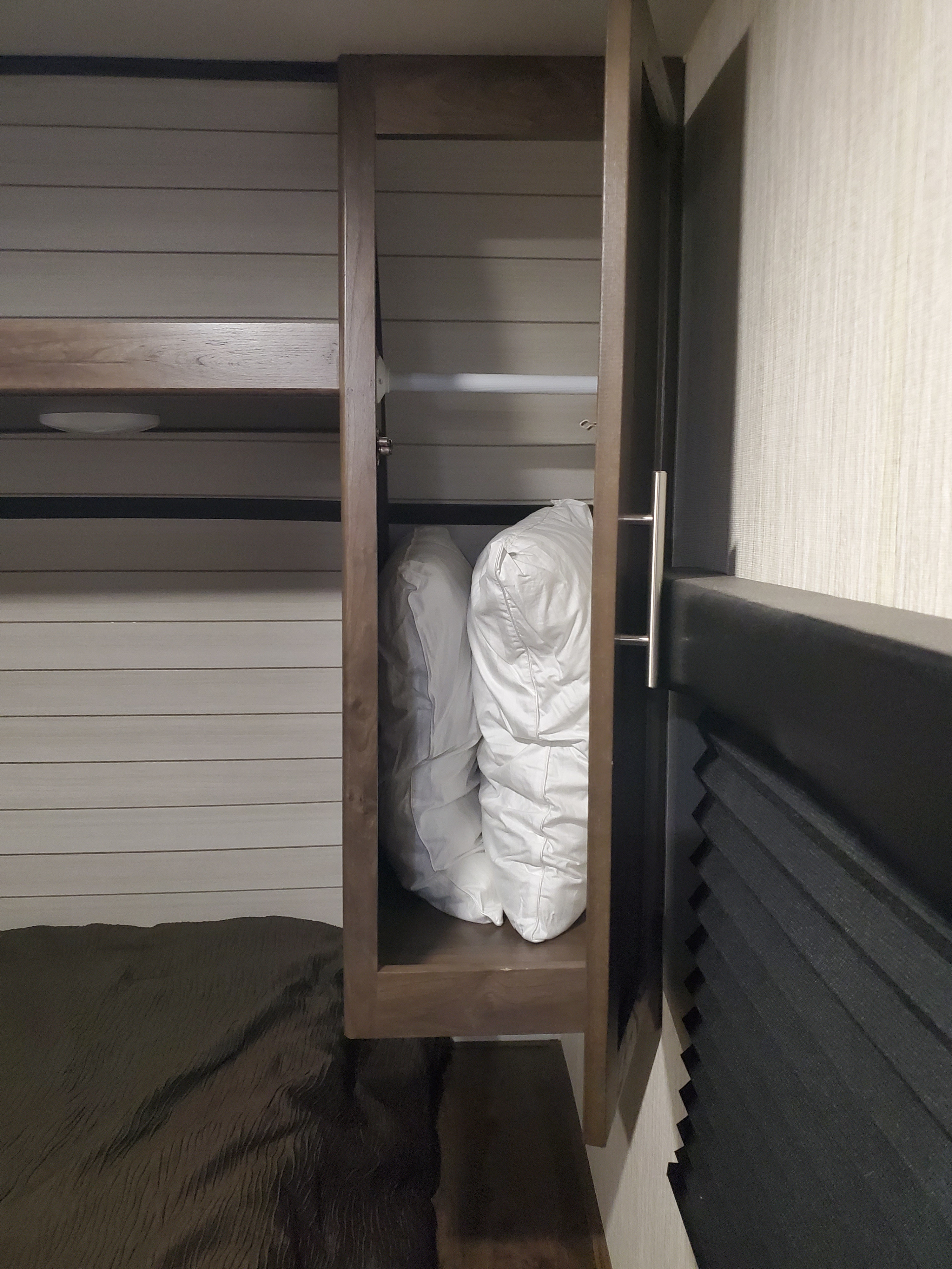 Inside an RV rental: a compact bedroom with an open cabinet storing pillows, dark bedding, and a window blind.. Other Other 2019