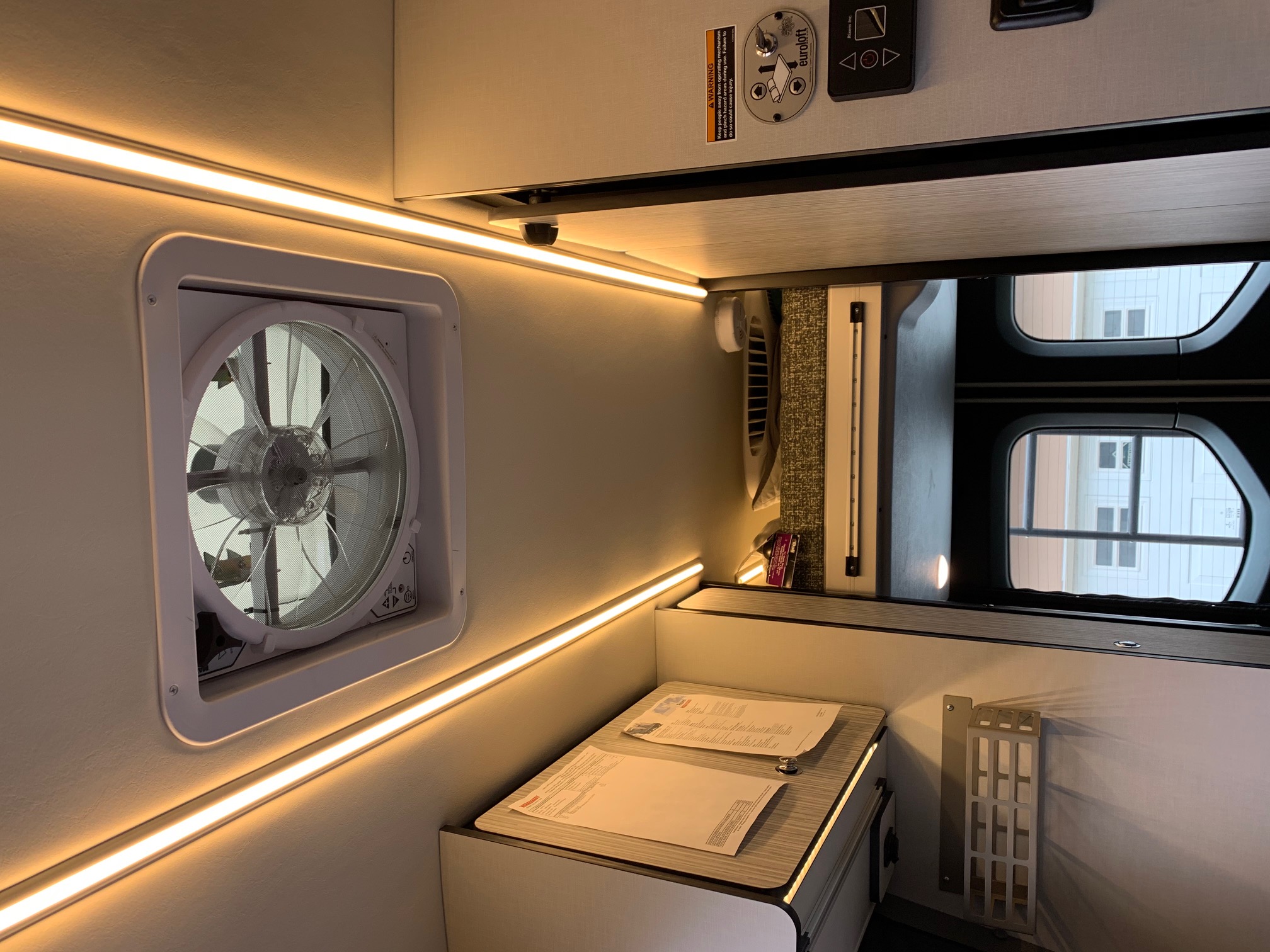 Inside a cozy RV rental: warm LED lighting, ceiling fan, and a desk area. A mirror shows the outside world.. Winnebago Revel 2020