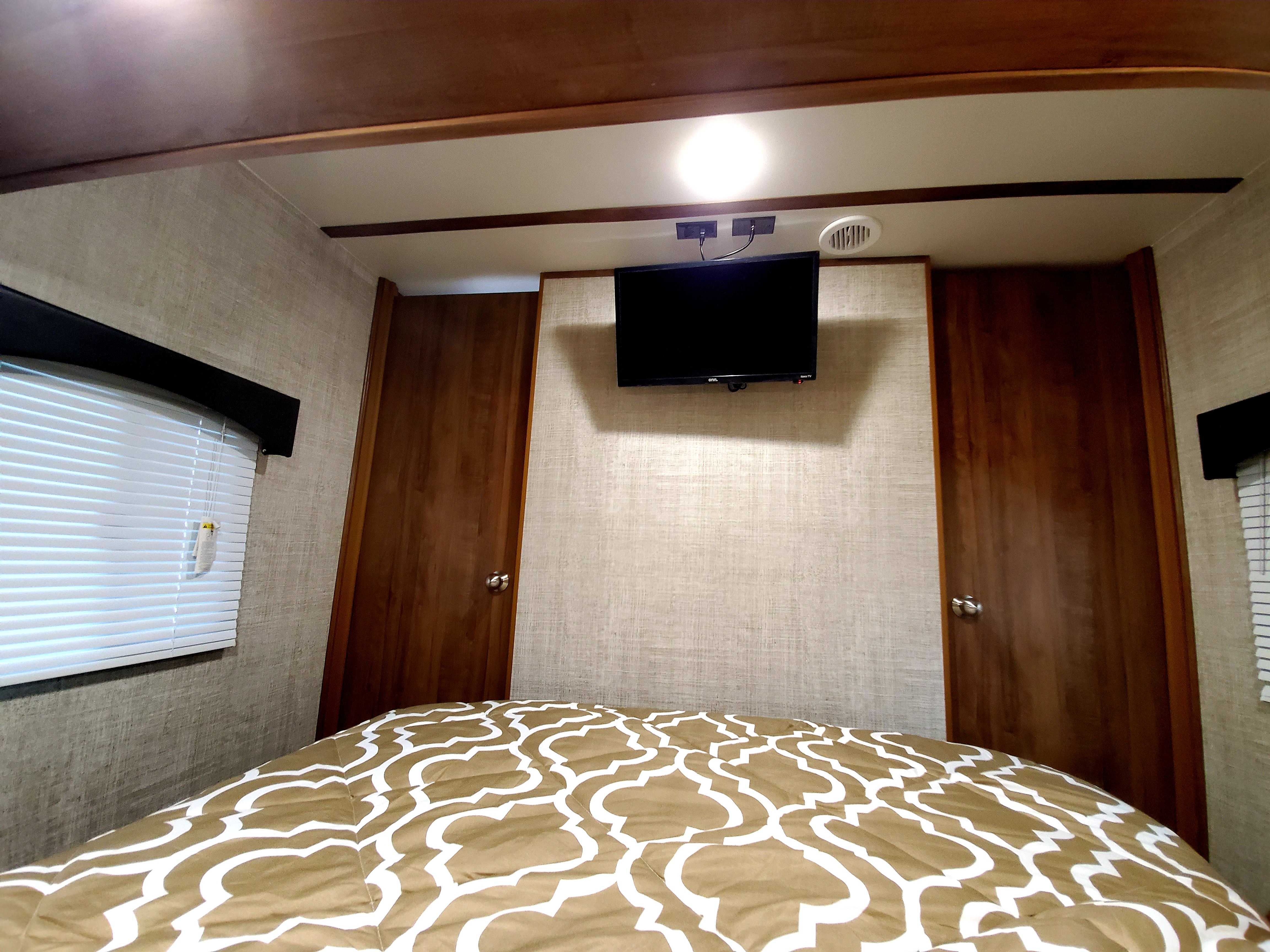 RV rental bedroom with TV above the bed, window with blinds, and two wooden doors, offering a compact interior view.. Gulf Stream 25BHS 2020