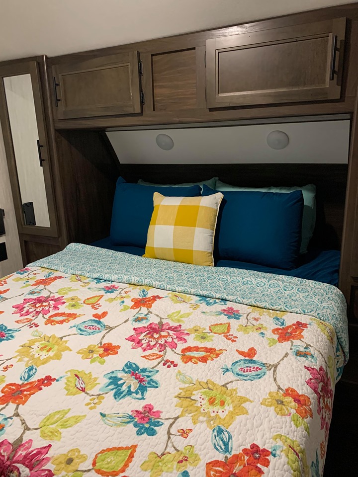 Inside an RV rental: a bright floral quilt covers a bed with blue pillows and overhead cabinets.. Forest River Cherokee Grey Wolf 2021
