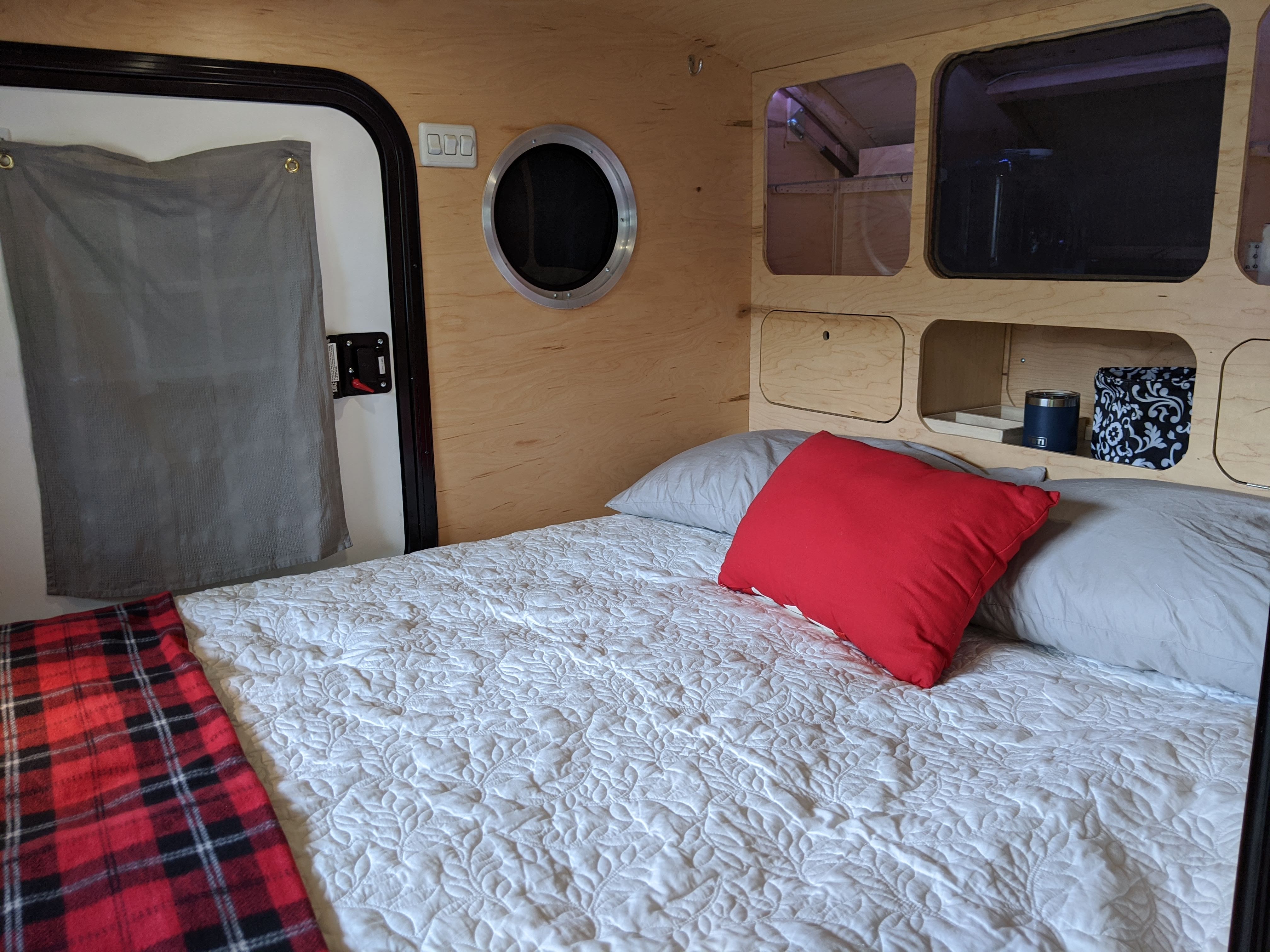 Inside a cozy rv rental: A comfy bed with white quilt, red pillow, built-in wood storage, and porthole window.. Custom Teardrop Camper 2019