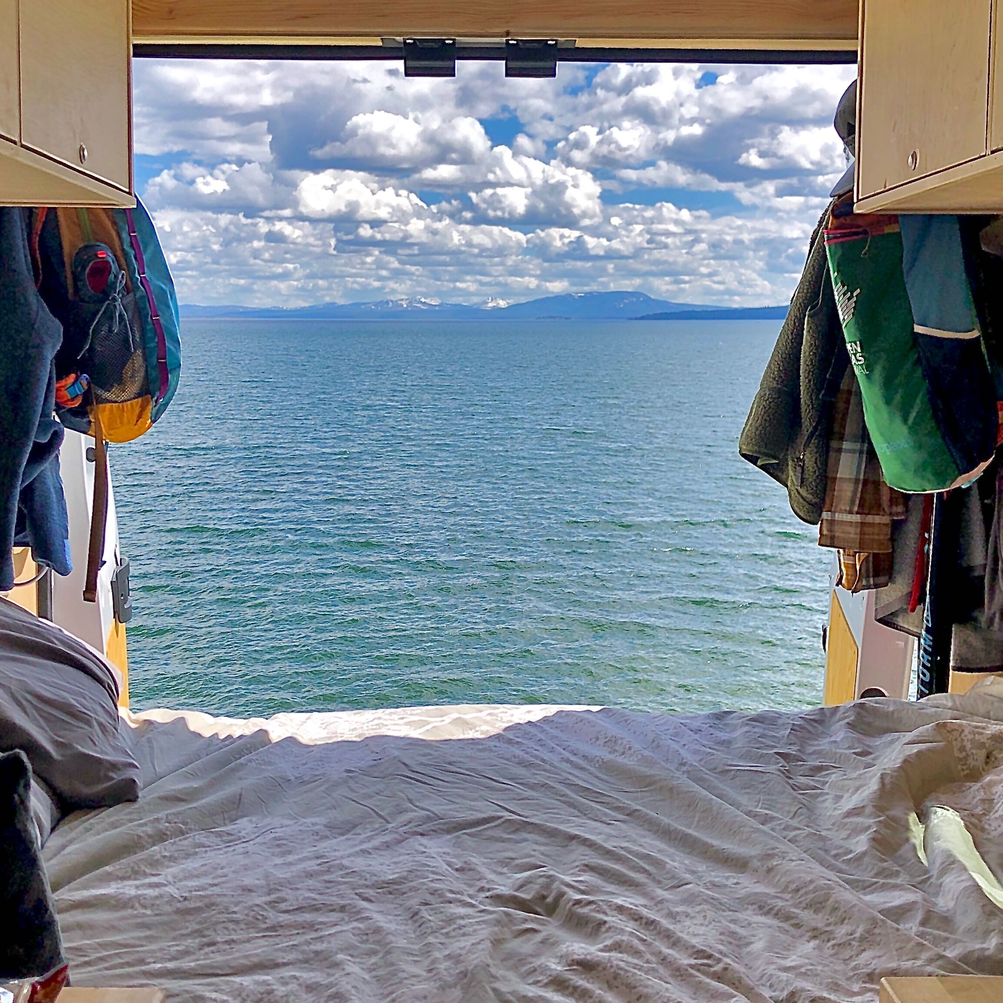 A cozy bed inside a camper van overlooks a calm lake, distant snowy mountains, and cloudy sky. Perfect RV rental escape!. RAM Promaster 1500 2017