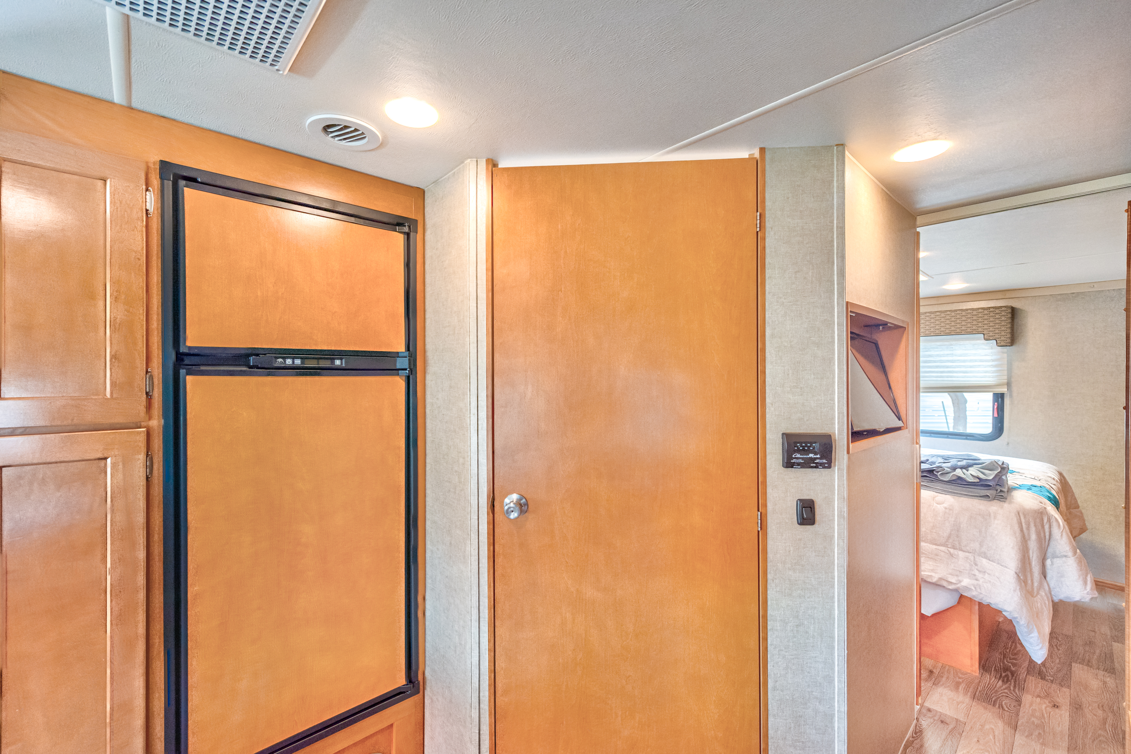 View of an RV rental's interior: wood-paneled refrigerator, main door, control panel, and a glimpse of the bedroom with a bed and window.. Winnebago Winnebago 2019