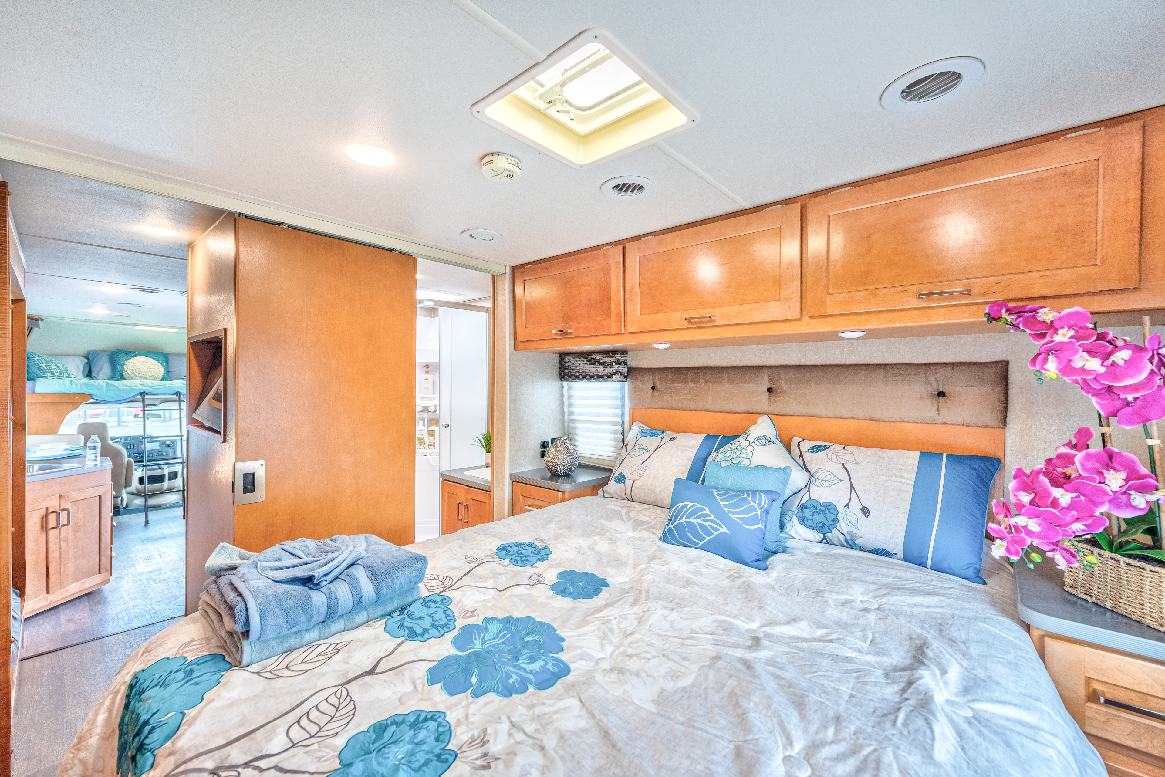 RV rental bedroom: cozy bed with blue floral bedding, overhead cabinets, and skylight. Sliding doors reveal kitchen/bunks.. Winnebago Winnebago 2019
