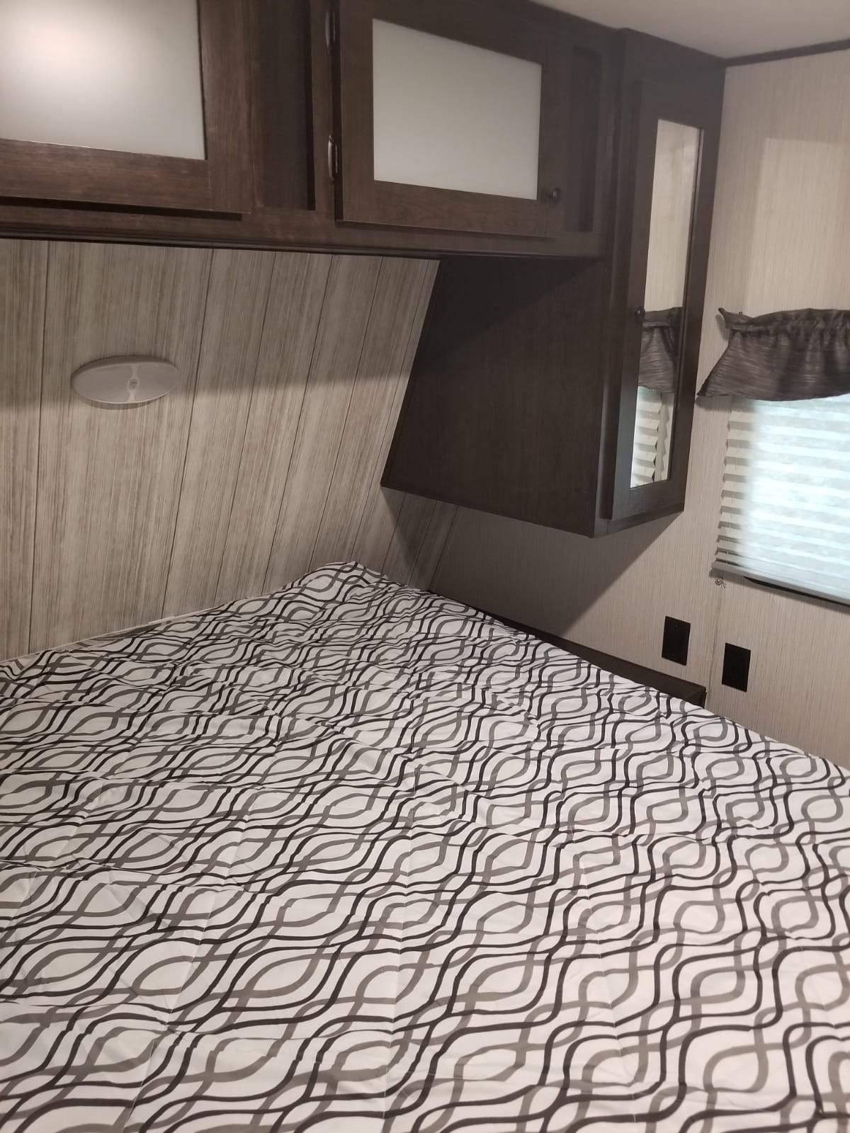 Inside an RV rental bedroom: cozy bed with wavy pattern, dark wood cabinets, and window with blinds.. Heartland Pioneer 2020