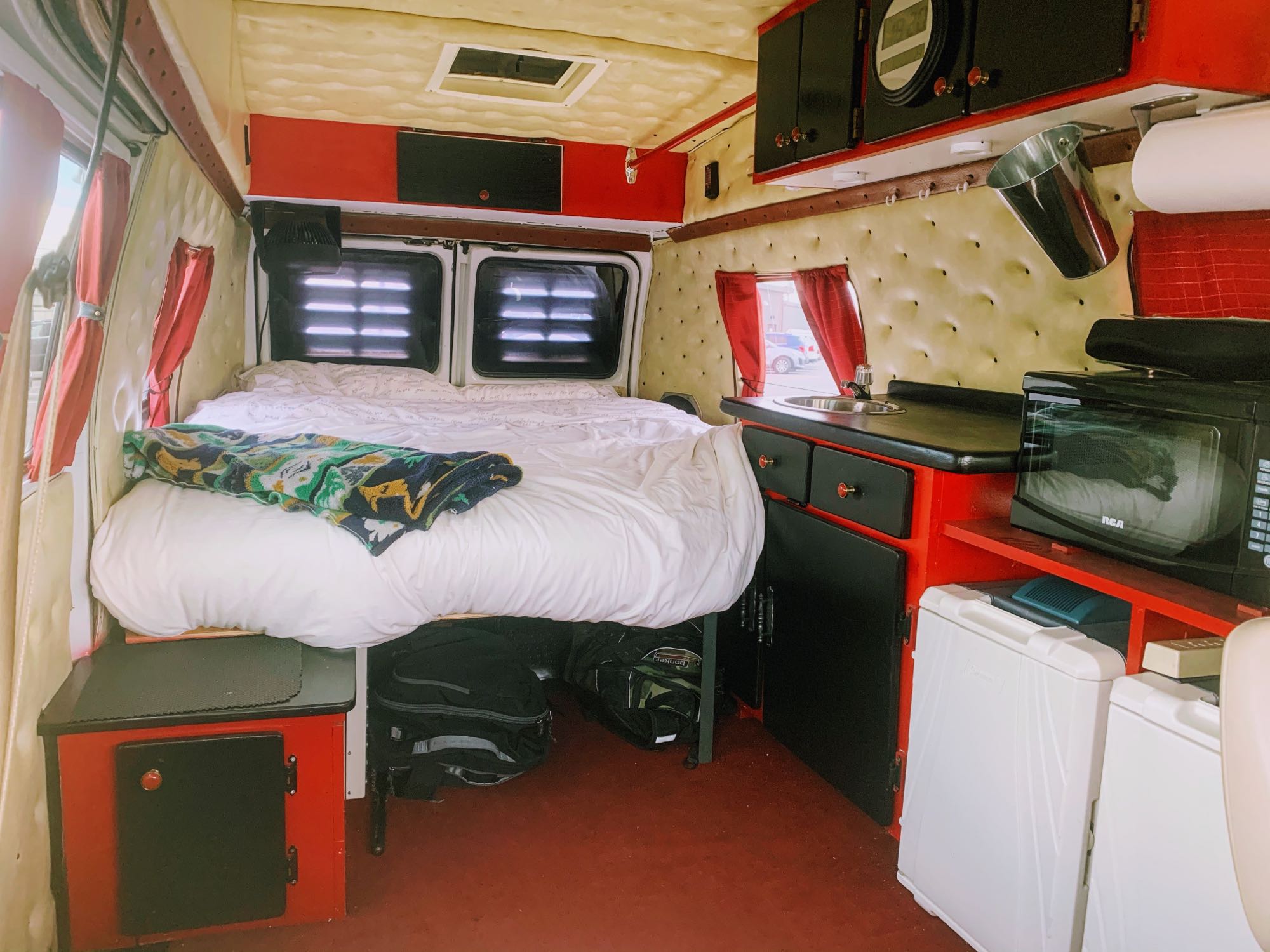 Explore this cozy campervan RV rental interior featuring a large bed, compact kitchen with sink, microwave, and fridge, all with stylish red and black accents.. Chevrolet G20 1983