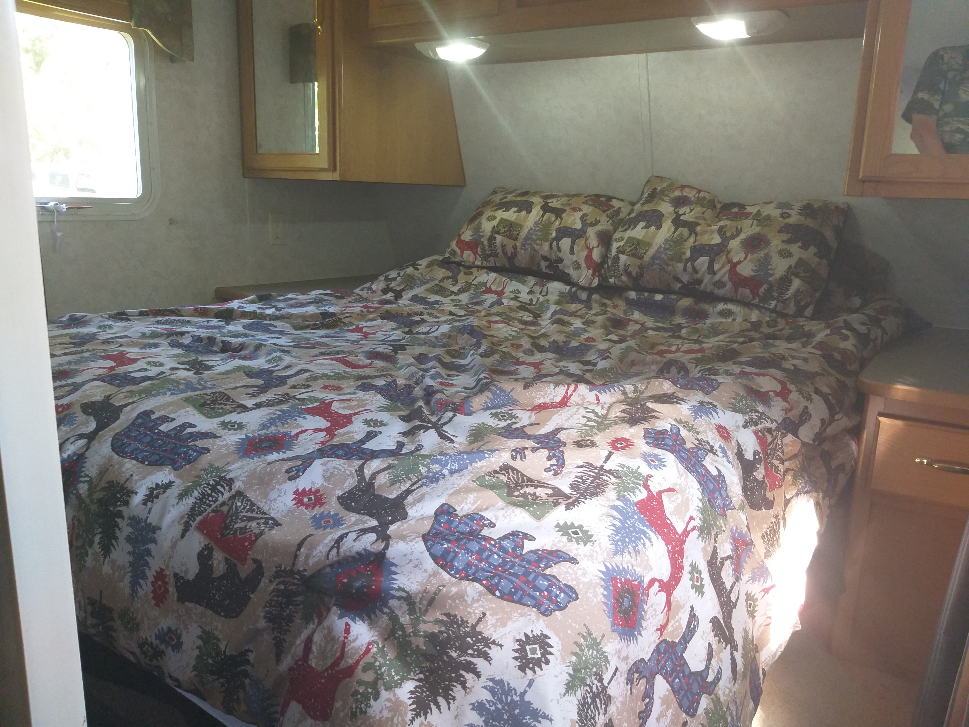 Cozy RV rental bedroom featuring a full bed with rustic deer and bear patterned bedding, overhead cabinets, and a side window.. Layton Layton Trailer 2007