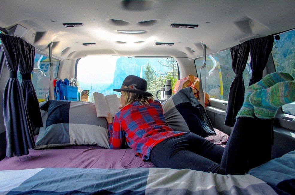 Cozy RV rental adventure: Person reads a book on a comfy bed inside a camper van, enjoying nature views.. Ford Econoline 150 2006