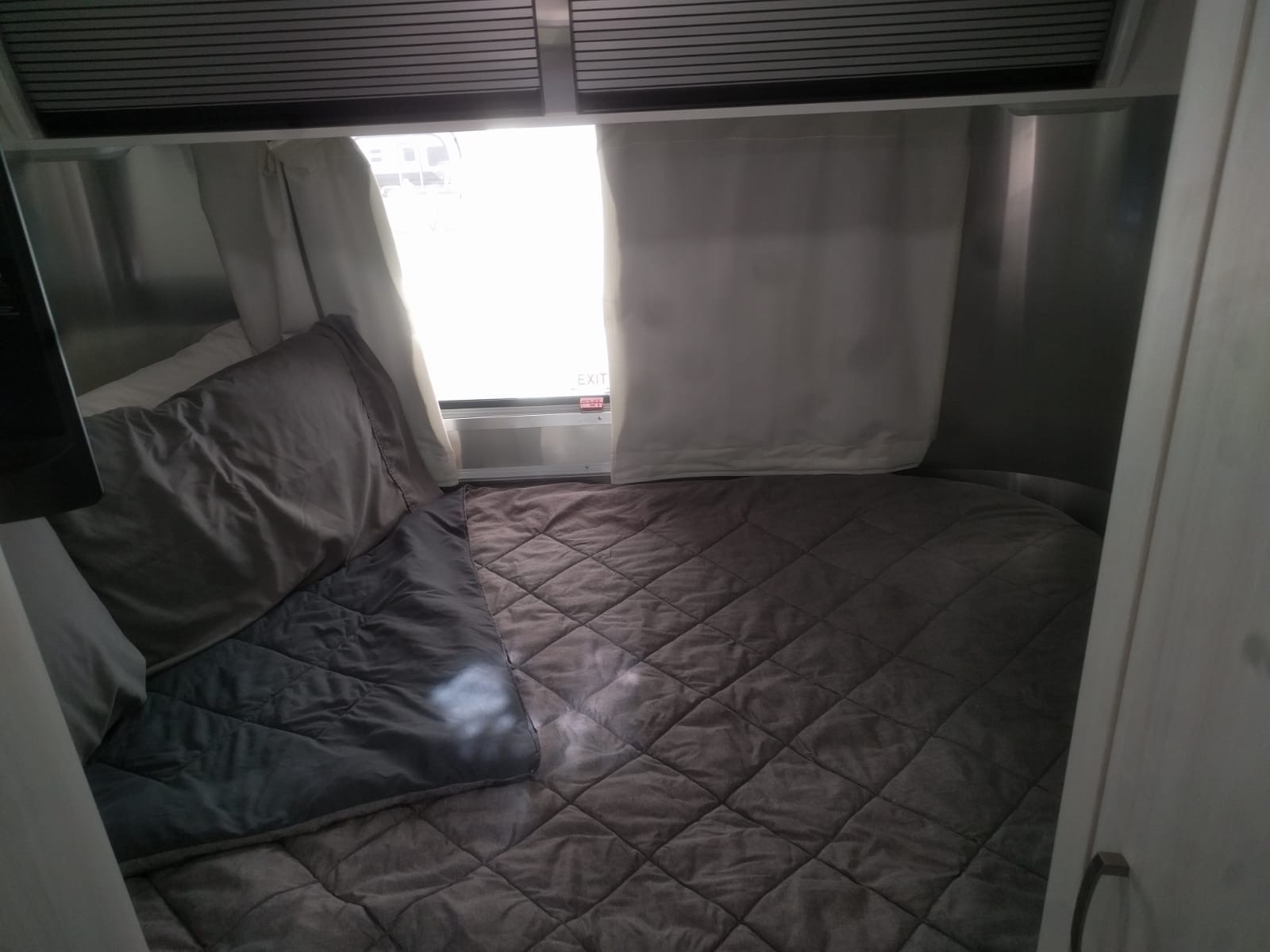 Cozy RV rental bedroom with a plush quilted bed, pillows, and a bright window featuring an "EXIT" sign.. Airstream Sport 2019