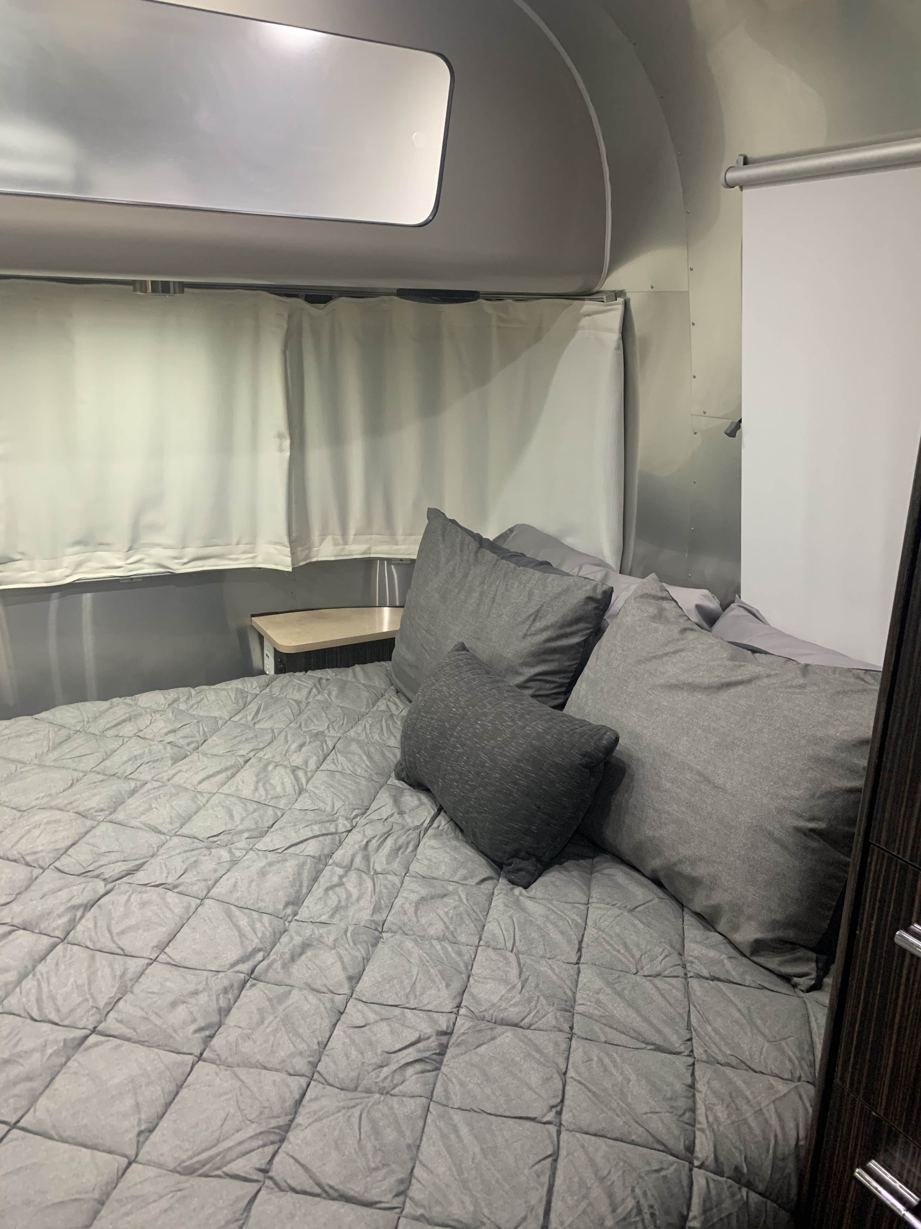 Inside an RV rental: a cozy bedroom with grey bed, pillows, and privacy curtains against polished aluminum walls. Bedside table.. Airstream International Signature 2019
