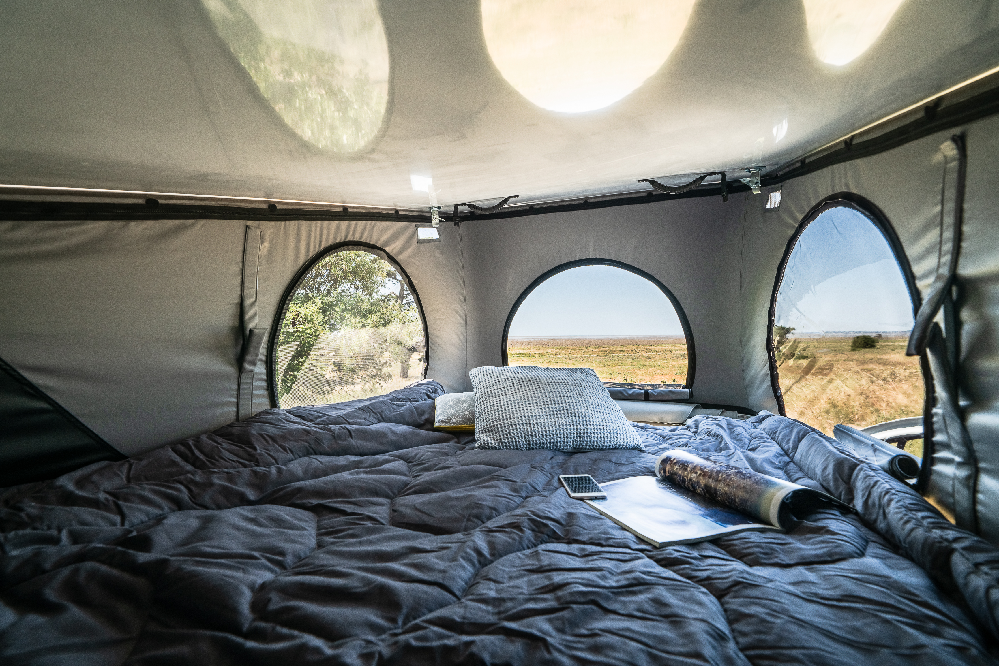 Cozy RV rental interior: a soft bed with grey bedding, smartphone & magazine. Arched windows offer stunning views of diverse landscapes.. Hanroad Trek 2023