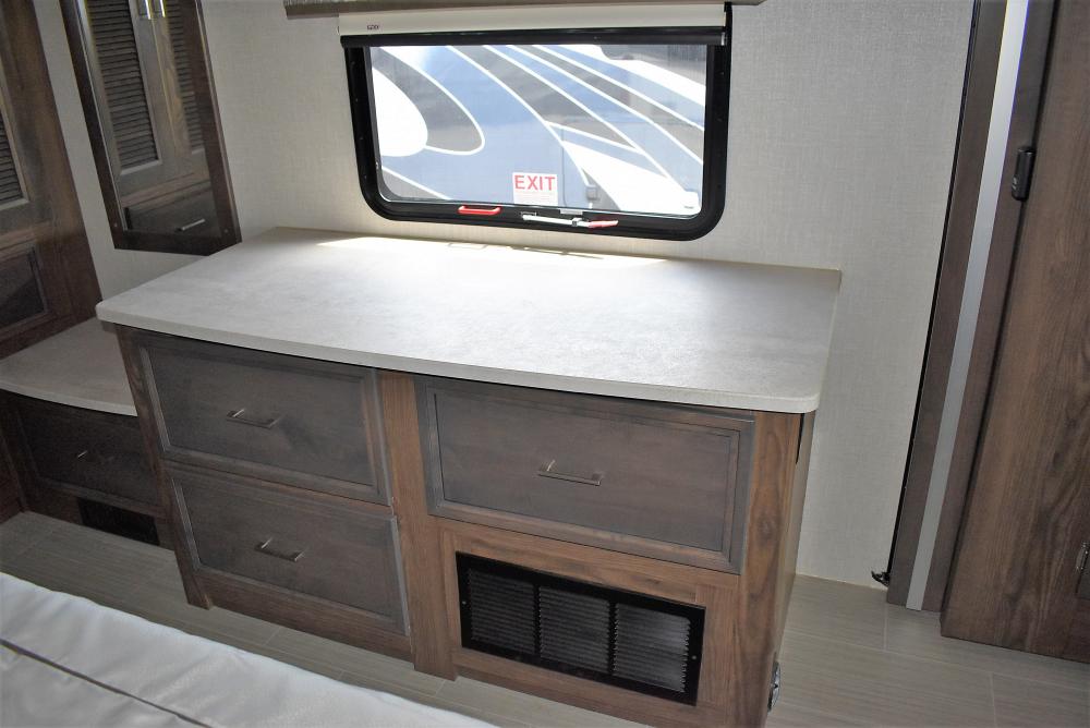 RV rental bedroom: A counter with storage drawers, a large window revealing an exterior 'EXIT' sign, and a padded bench.. Coachmen Sportscoach 2020