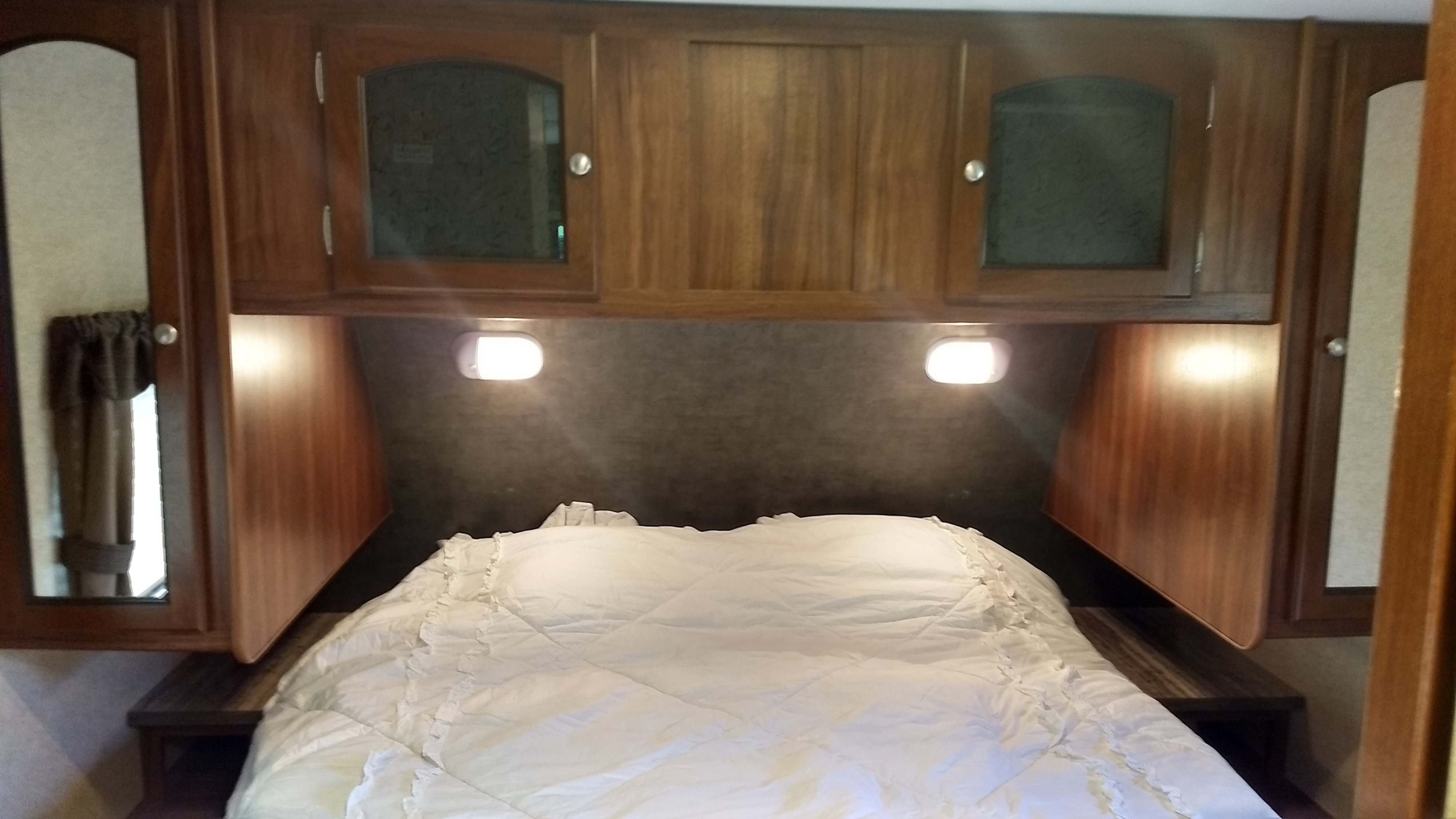Inside an RV rental bedroom: cozy bed with white comforter, overhead wood cabinets, and reading lights.. Heartland North Trail 2015