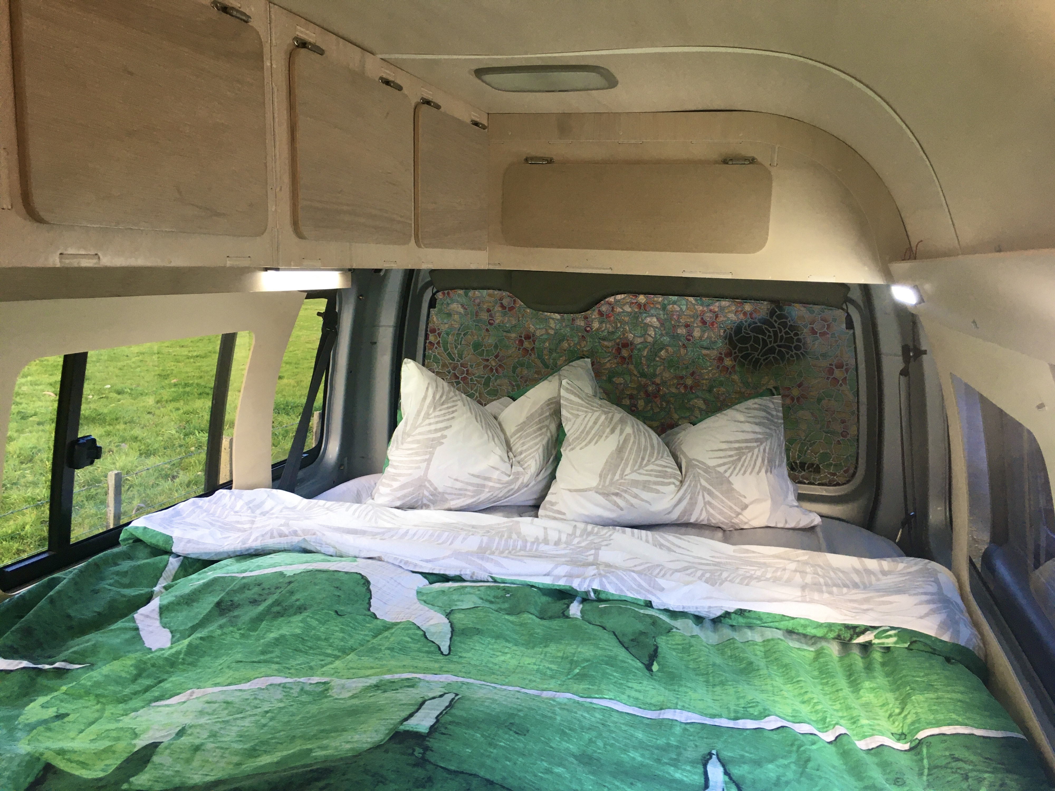 Inside a comfortable RV rental campervan, featuring a cozy bed with green bedding, overhead storage, and a field view.. Toyota Hiace 2006