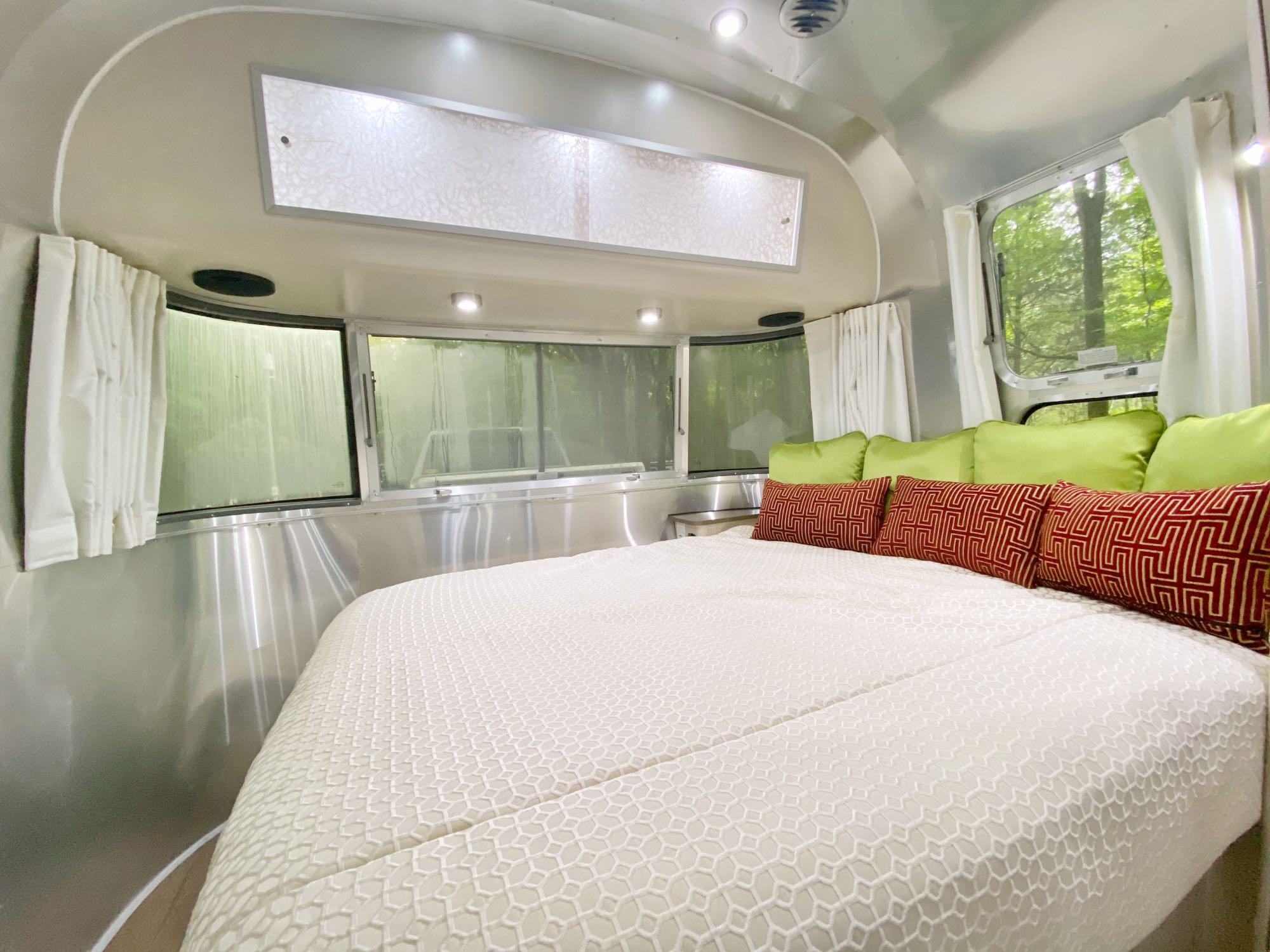 Modern Airstream RV rental bedroom: comfy bed with white duvet, green & red pillows, and lush nature views from windows.. Airstream International 2019