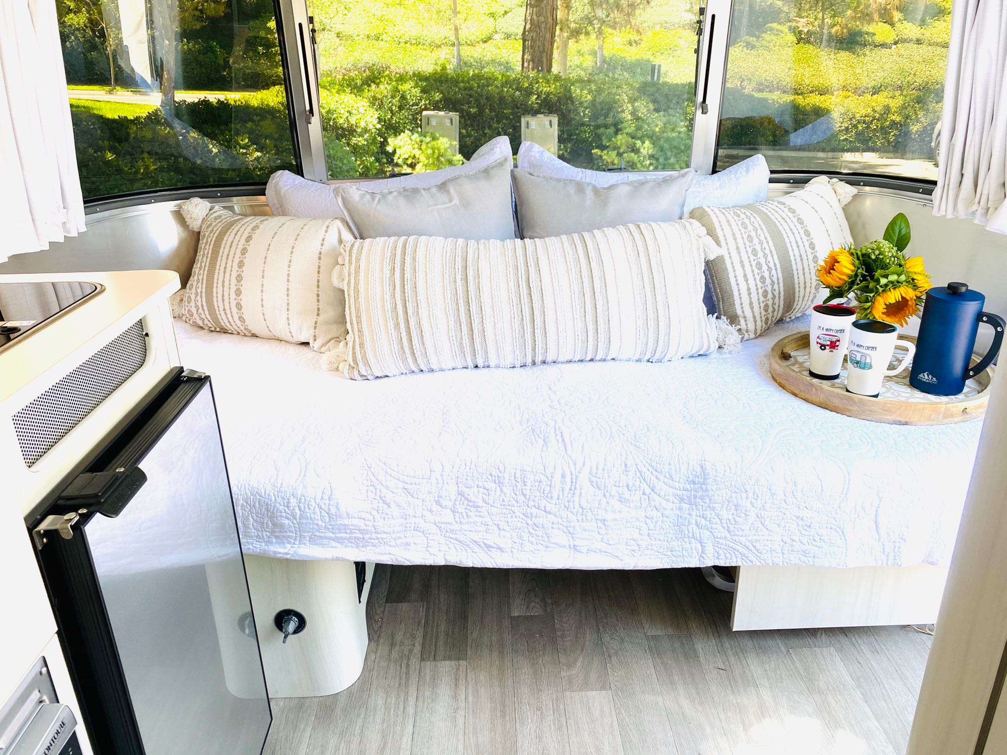 Relaxing RV rental interior: A comfy bed with decorative pillows & sunflowers, brightened by nature outside the large windows.. Airstream Bambi 2020