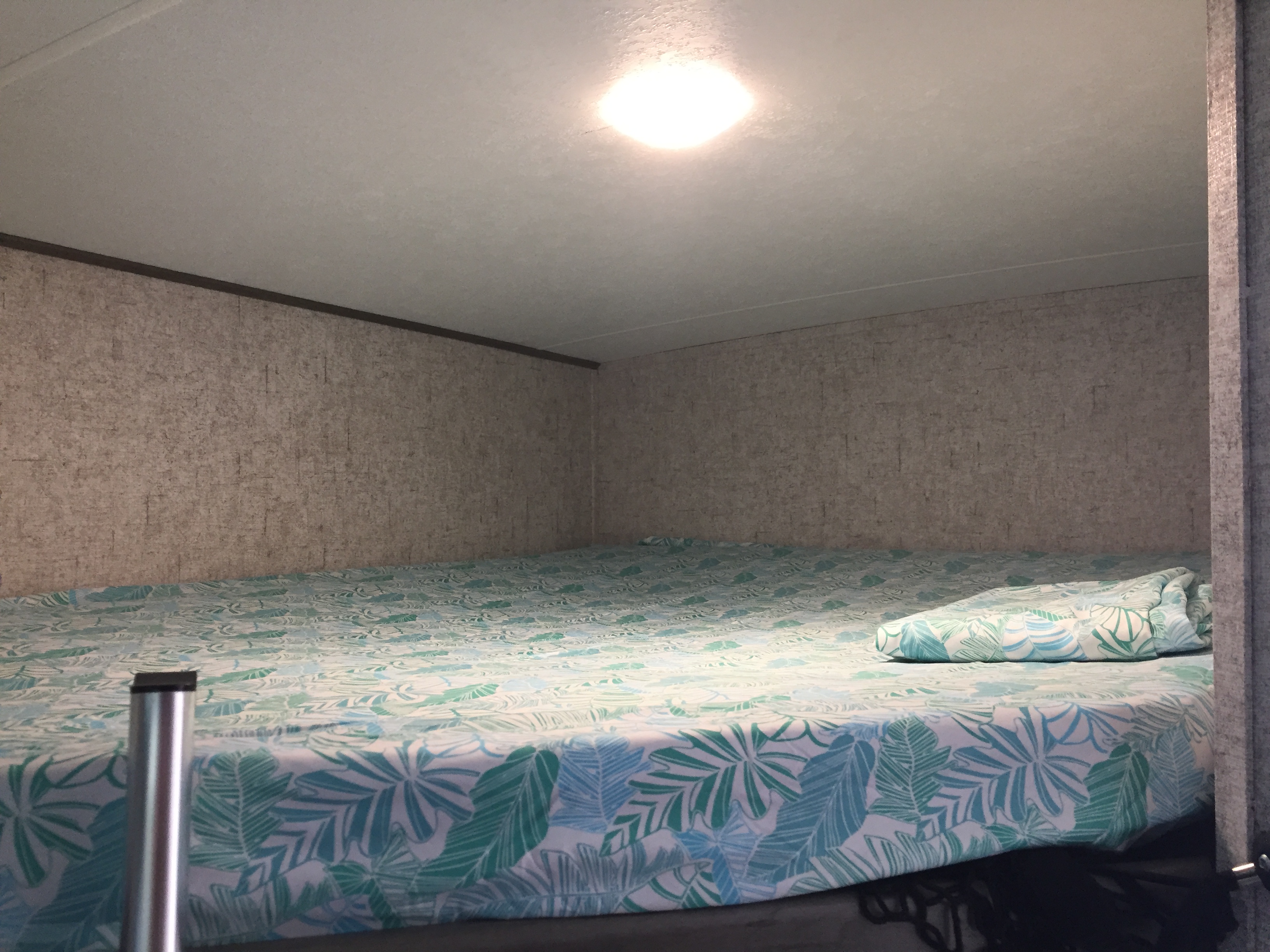 **RV rental** interior: a cozy bunk bed with blue & green tropical leaf sheets under a bright ceiling light. Textured walls.. Coachmen Apex 2021
