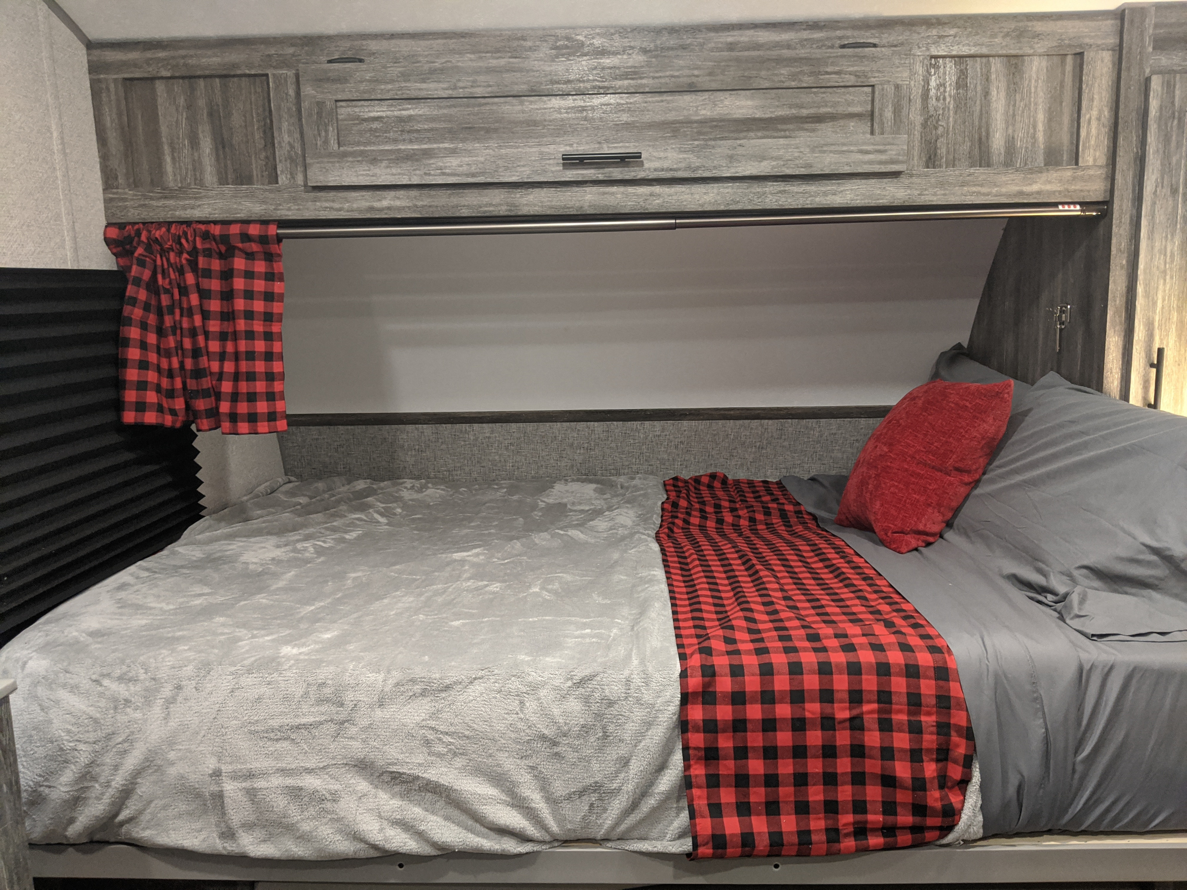 Comfortable RV rental bed with grey bedding, red plaid throw, black blinds, and distressed wood-look cabinets.. Forest River Wildwood 2021