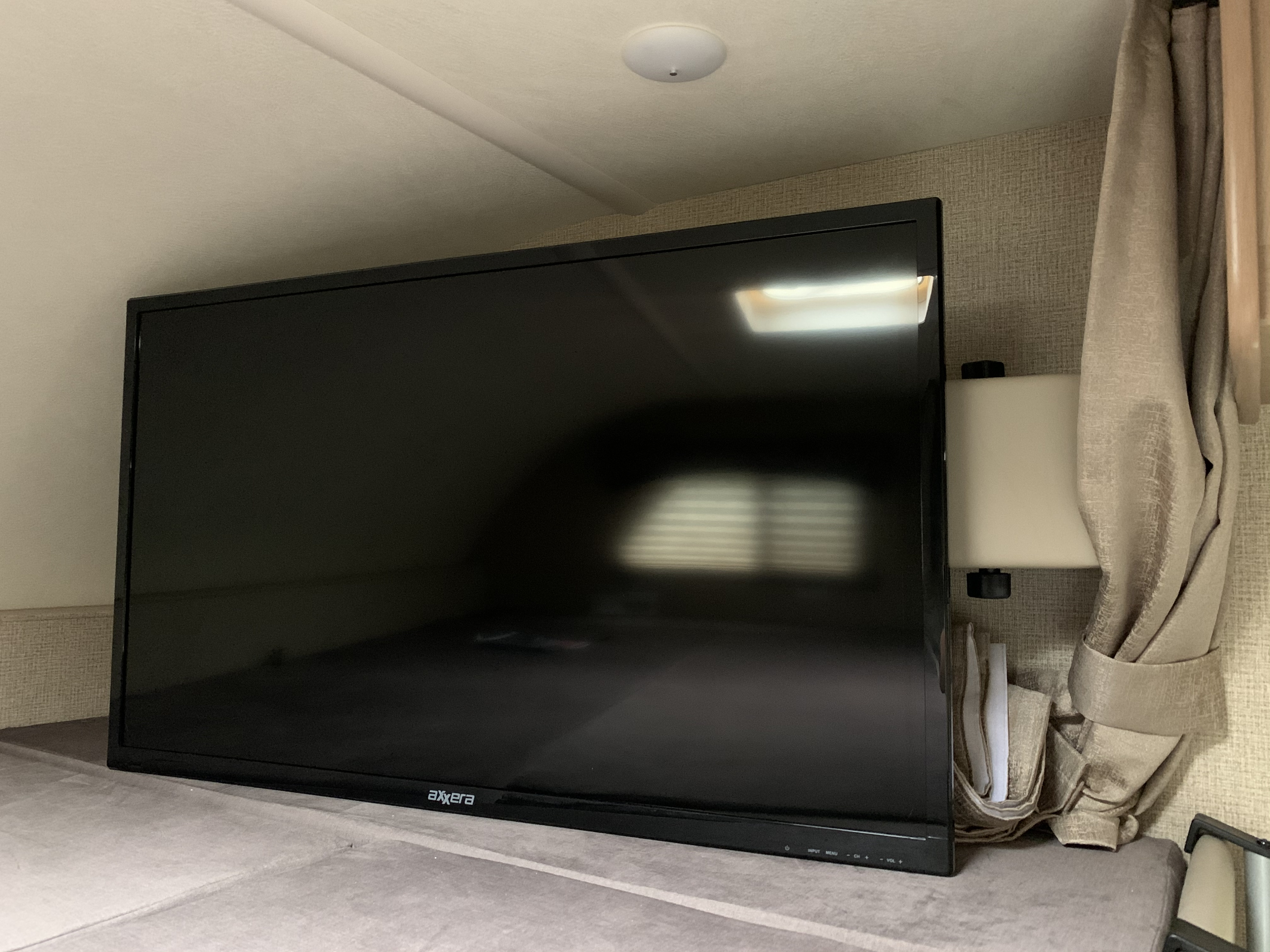 An Axxera flat-screen TV, part of an RV rental, rests on a cushioned surface, reflecting a light and blinds.. Thor Chateau 2019