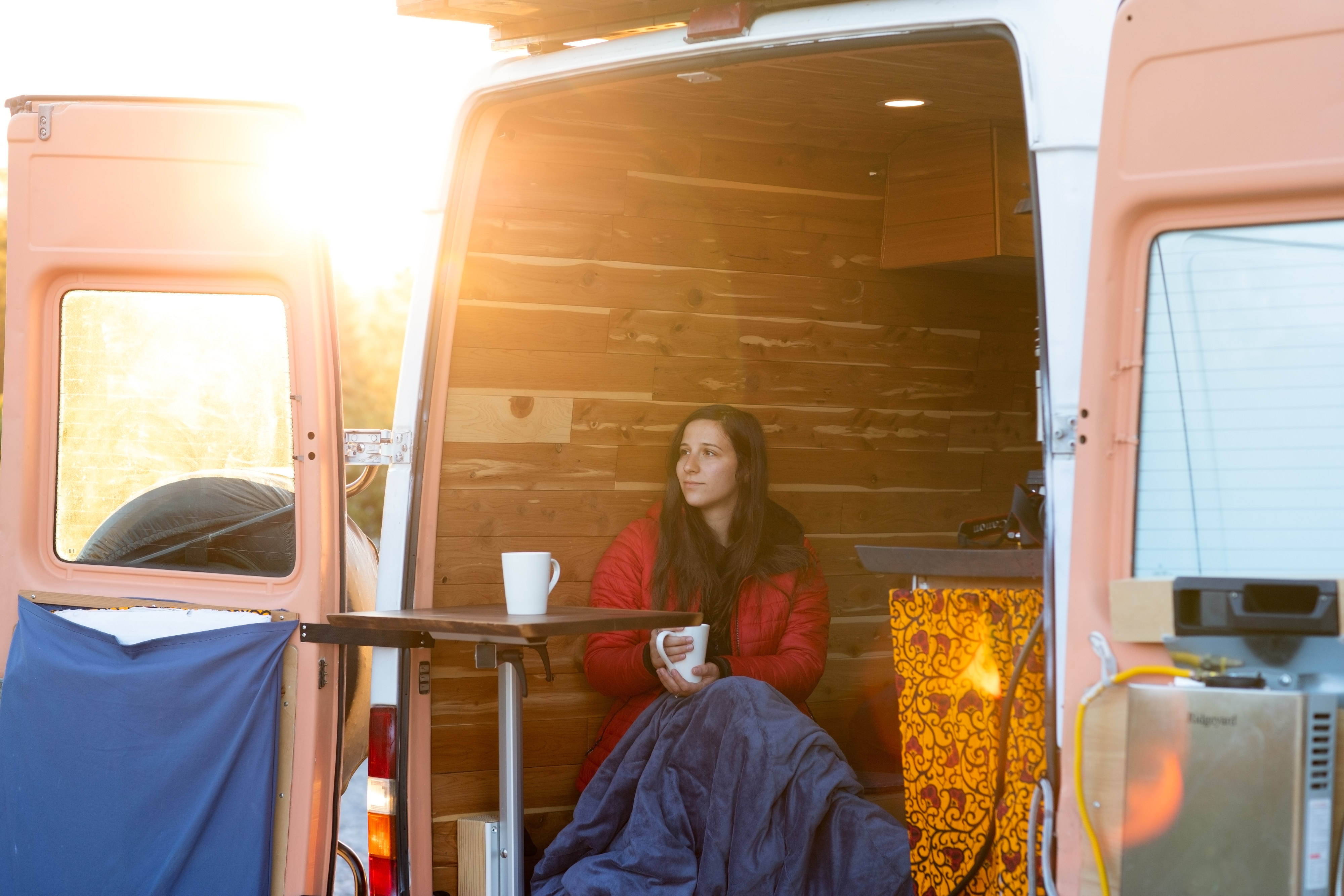 Woman bundled in blanket sips warm drink inside sunlit, wood-paneled RV rental van.. Dodge Sprinter 2004