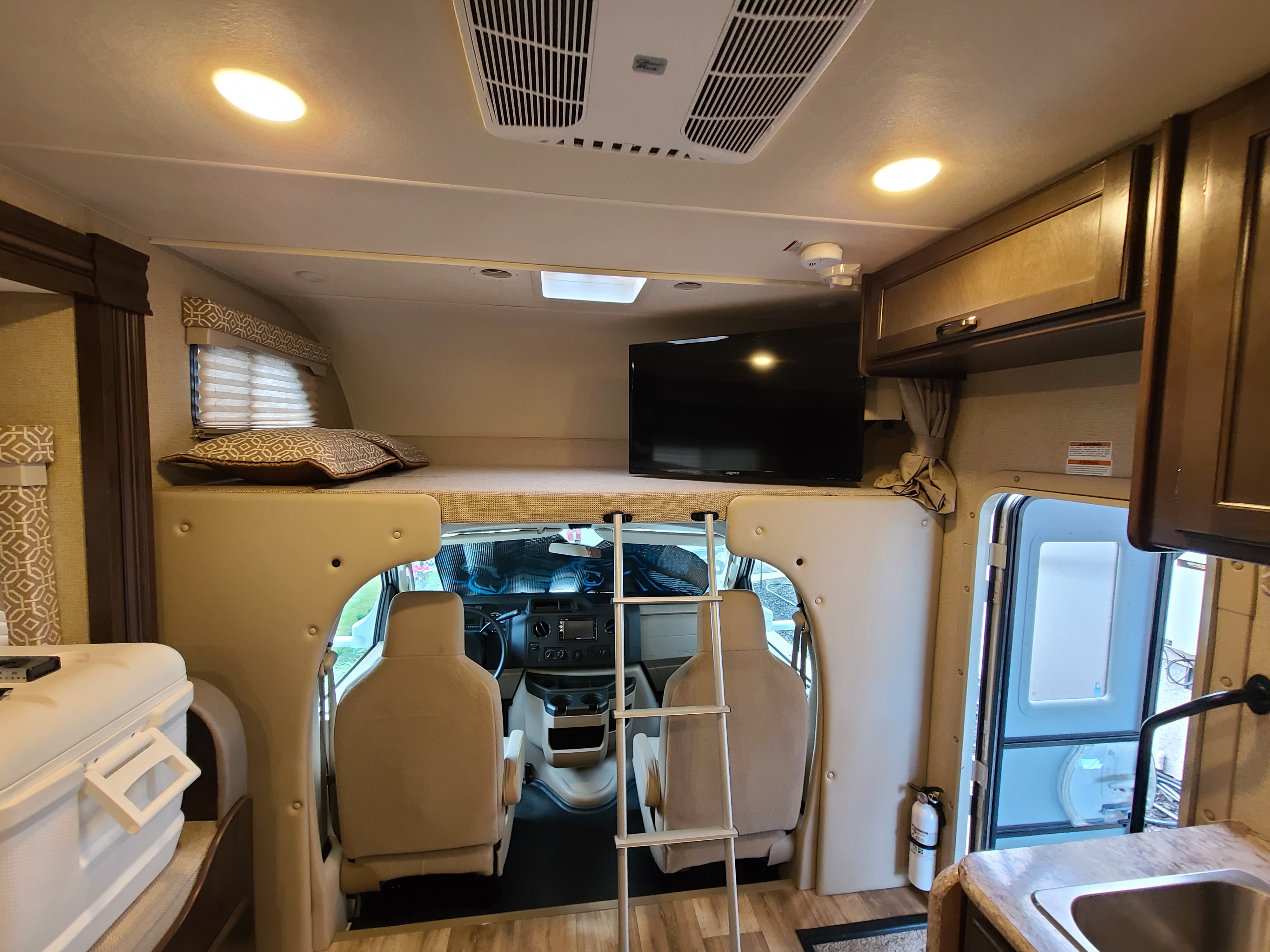 Inside an RV rental: a forward view showing the spacious overhead bunk with TV, ladder, driver's cockpit, and entrance.. Thor Freedom Elite 2019
