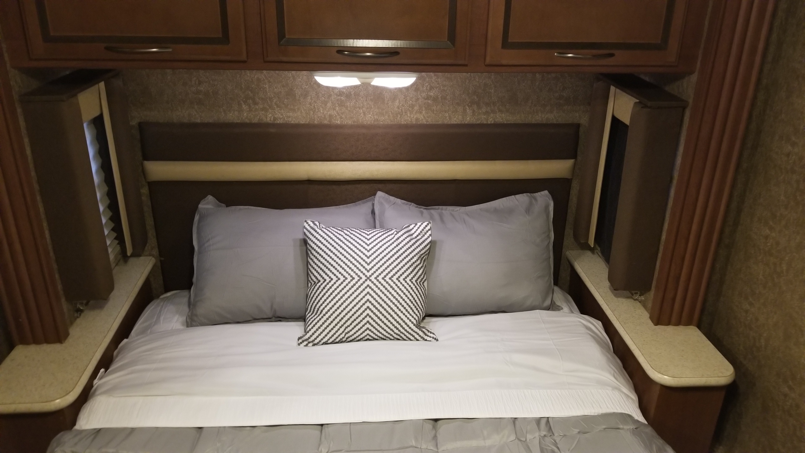 Cozy RV rental bedroom with a neatly made bed, grey pillows, patterned cushion, overhead cabinets, and side shelves.. Thor Motor Coach A.C.E 2013