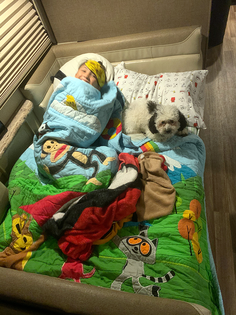 Enjoying an RV rental: A child smiles cozily in bed with a white dog. Comfy blankets & pillows for travel adventures.. Thor Motor Coach A.C.E 2020