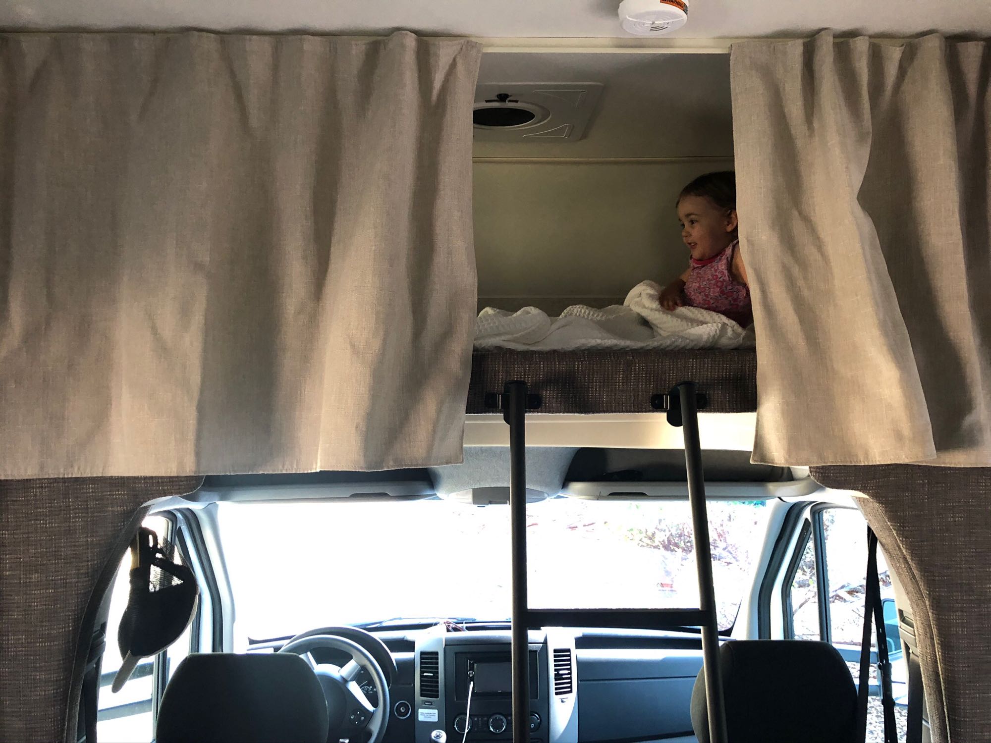 Inside an RV rental: A child rests comfortably in the overhead bunk, with the driver's cab visible below.. Winnebago Vita 2020