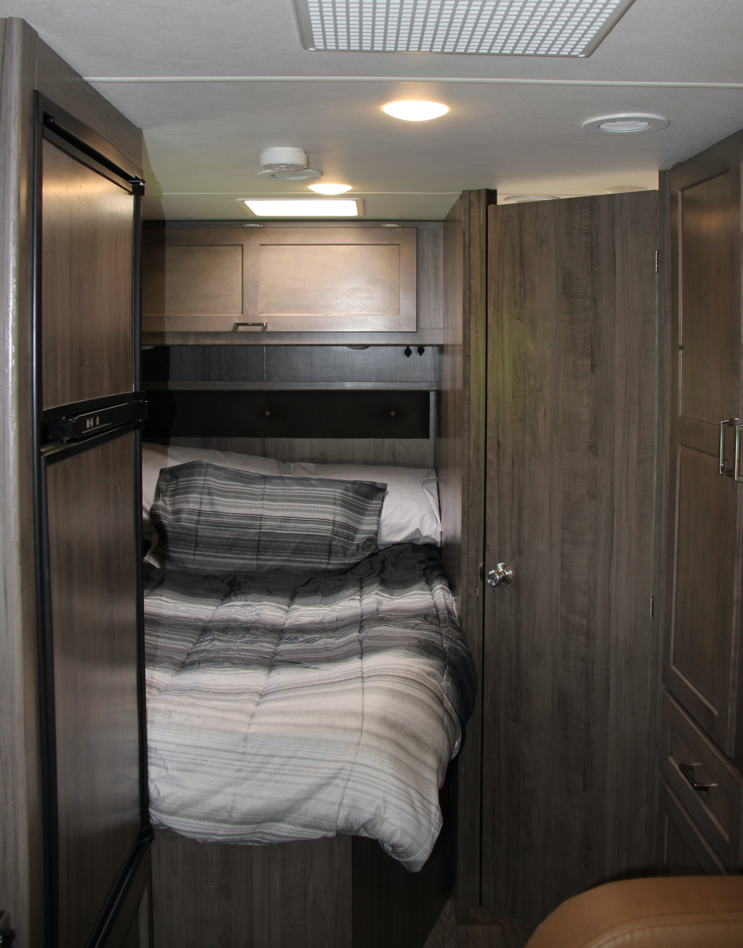 Compact RV rental bedroom featuring a striped bed, refrigerator, overhead cabinets, and ceiling fixtures.. Winnebago Minnie Winnie-Hook 2019