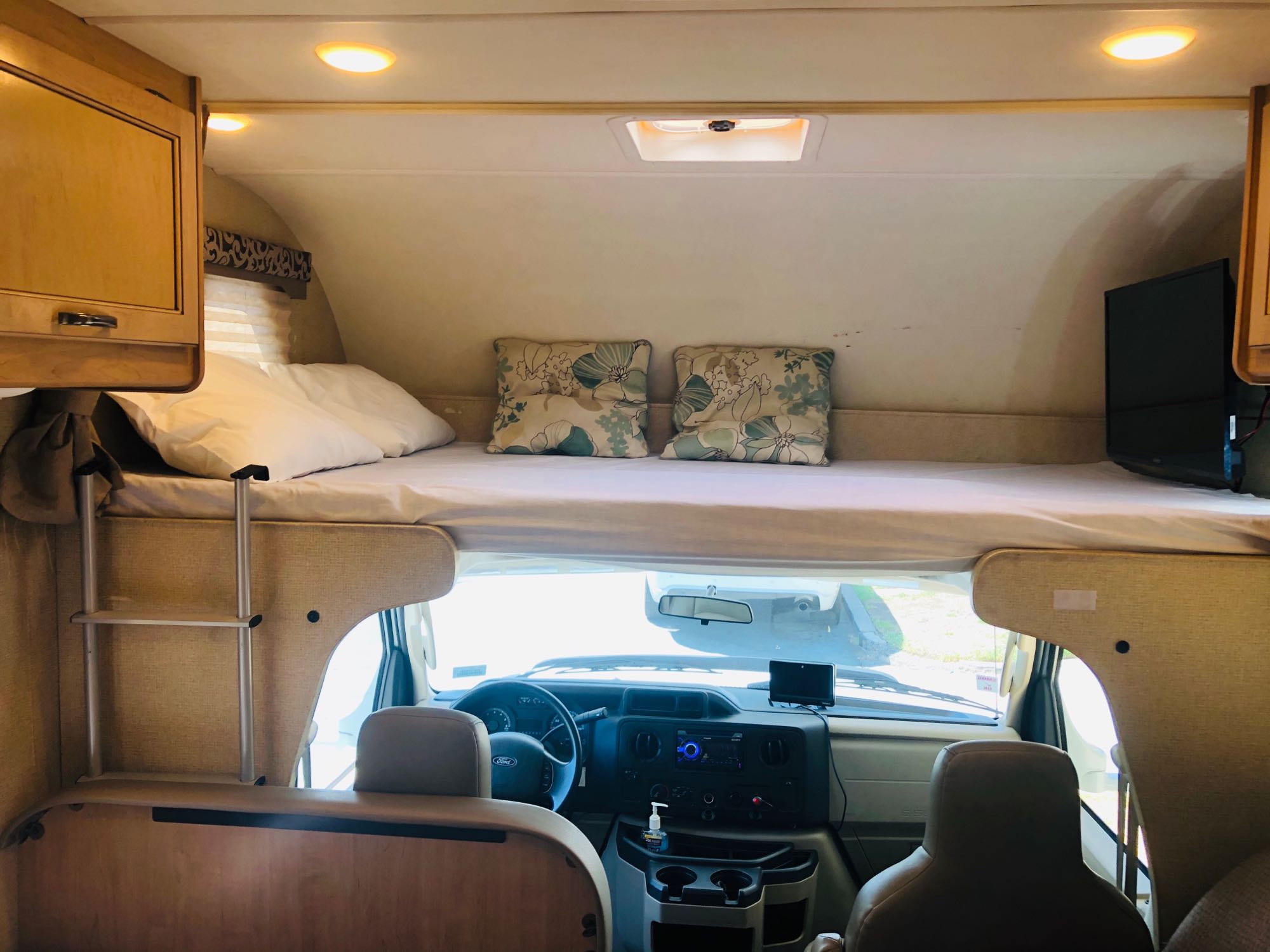 Cozy RV rental interior shows a large overhead bed, ladder, TV, and the driver's cockpit with a Ford steering wheel.. Thor Four Winds 2019