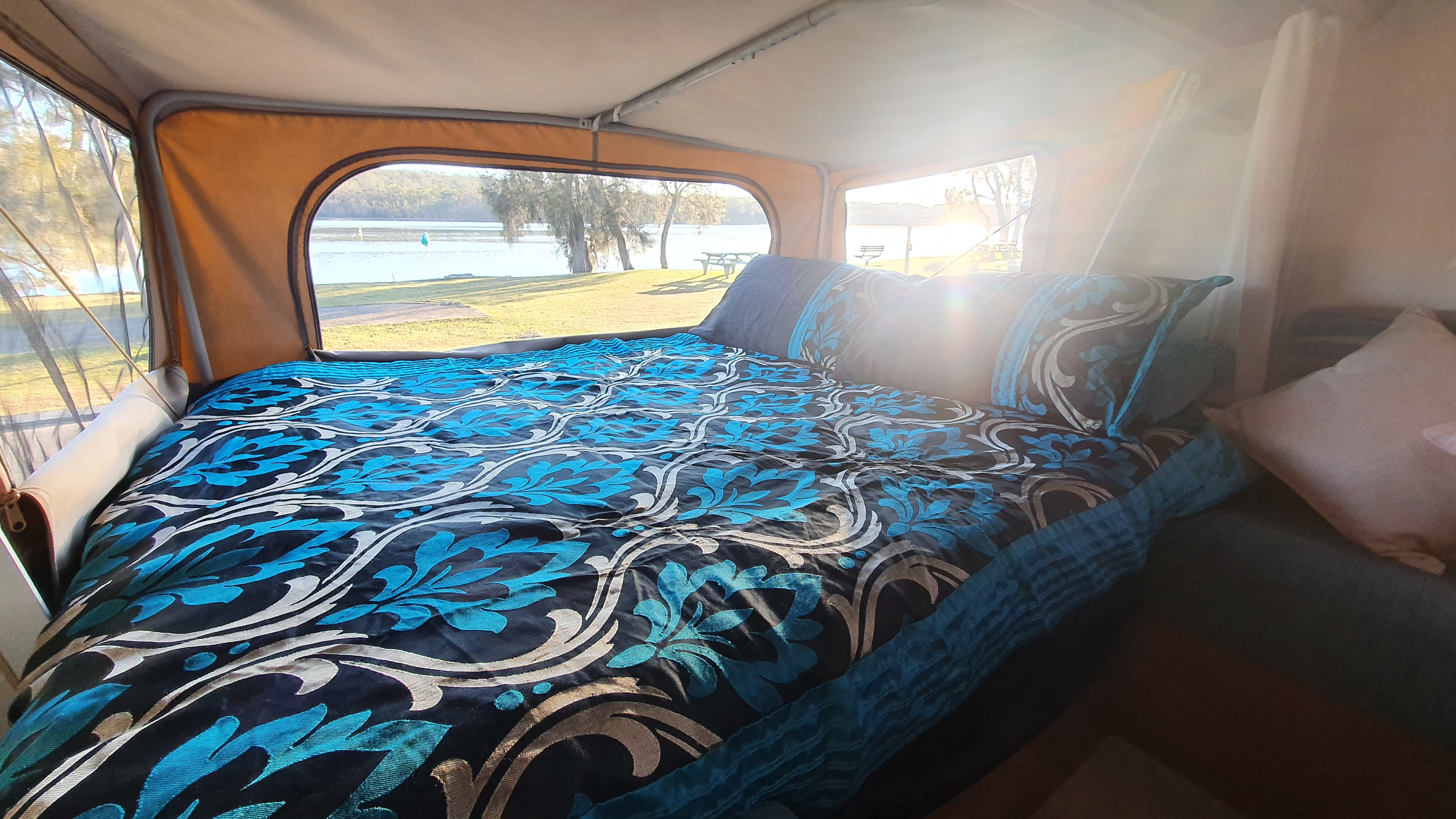 Inside a cozy RV rental pop-up camper with a patterned bed and pillows, overlooking a scenic lake.. jayco Expanda 2010