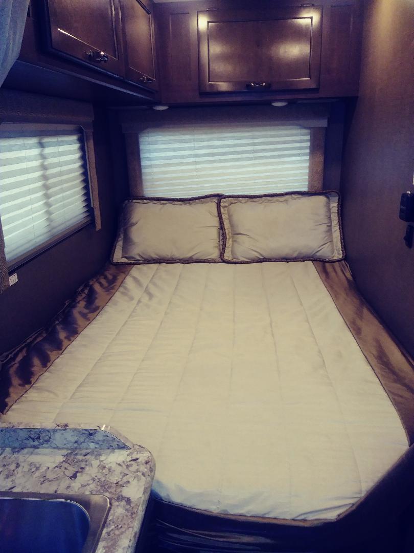 Inside an RV rental: a cozy bedroom with a neatly made bed, window blinds, and overhead cabinets. A compact sink is also visible.. Thor Freedom Elite 2018